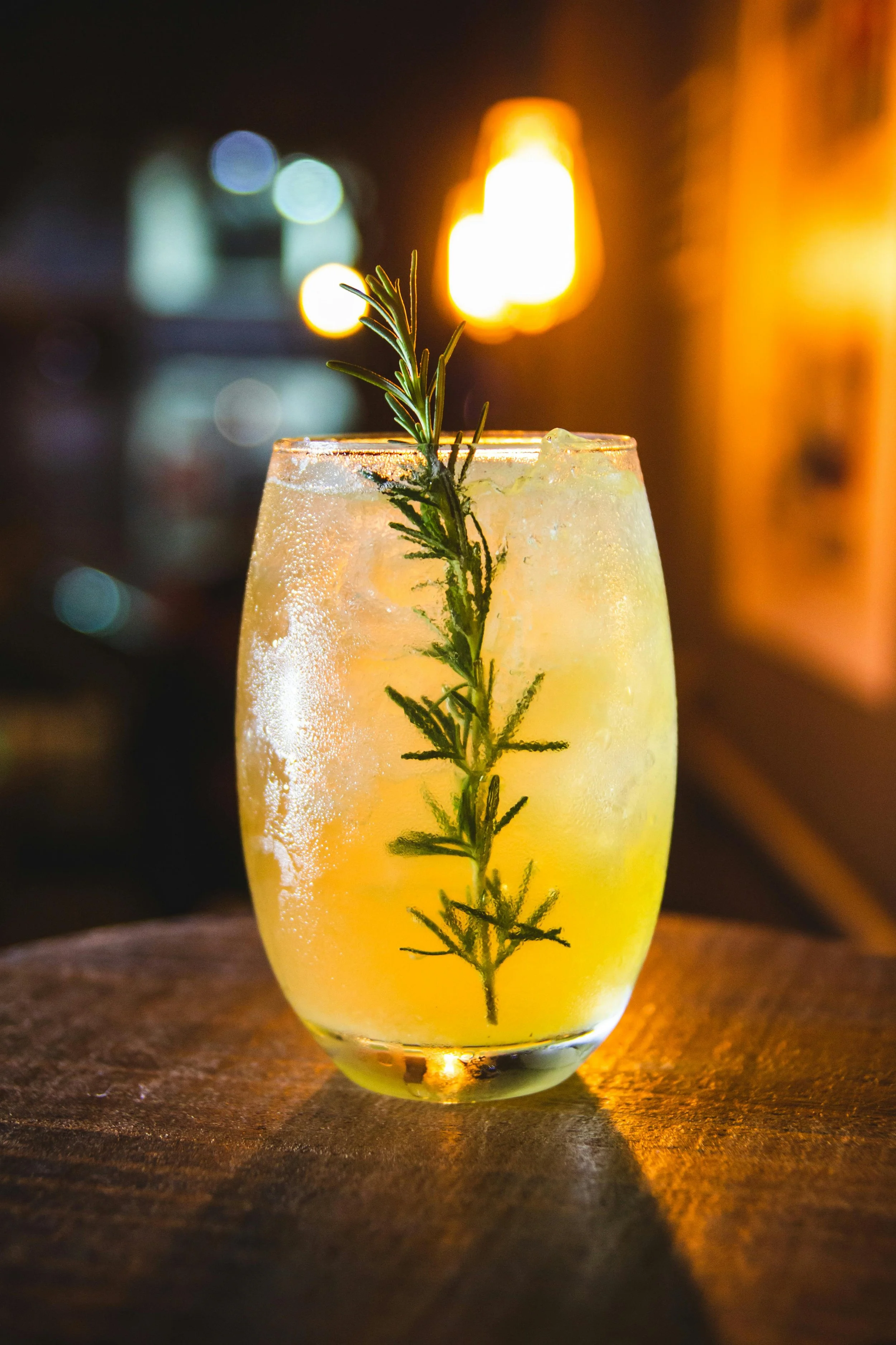 Close-up of a chilled cocktail glass with a yellow drink and a sprig of rosemary, set on a wooden table with a warm, dimly lit background.