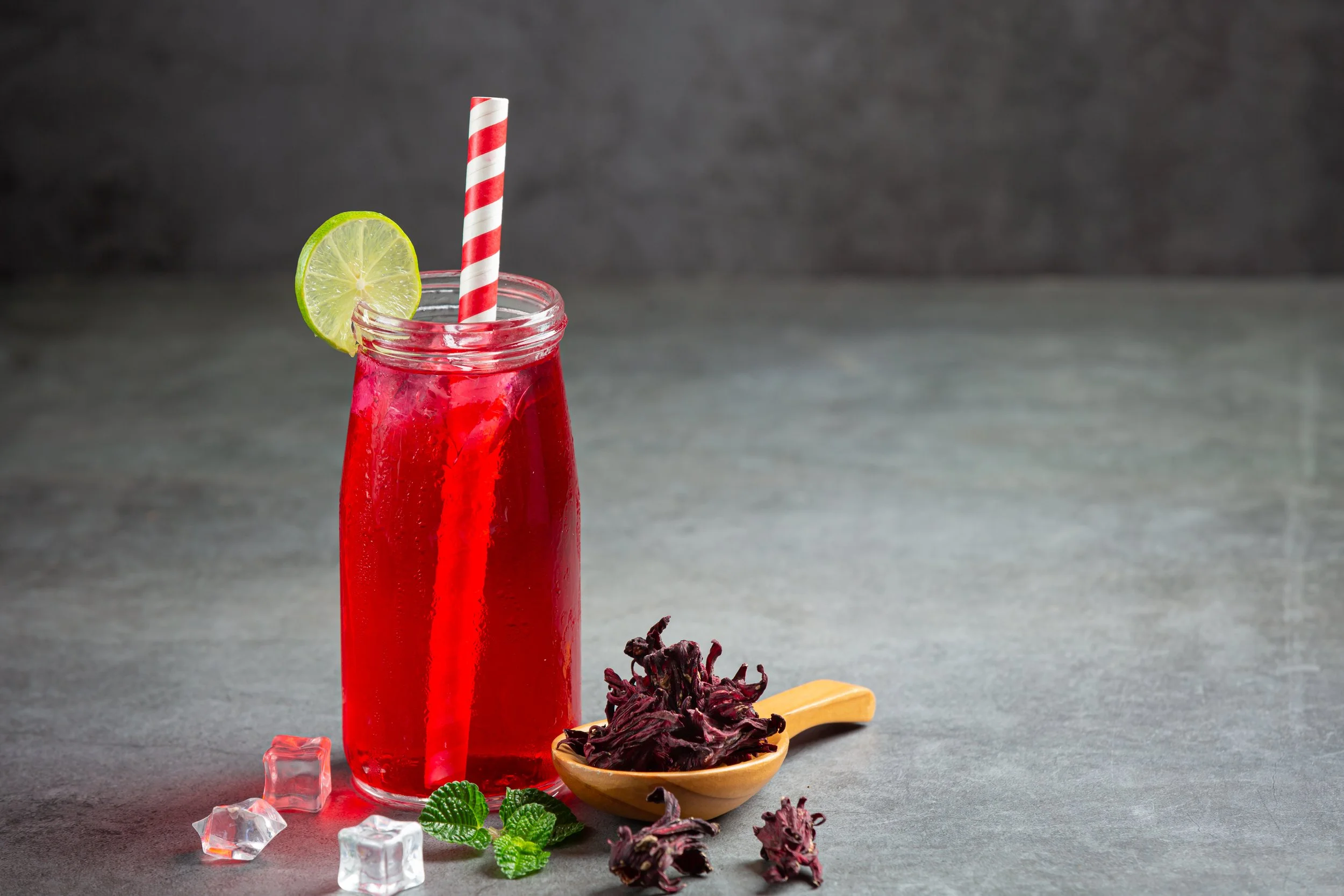 A glass of red hibiscus tea with ice, a striped straw, and a lime slice on the rim, surrounded by hibiscus flowers, mint leaves, and ice cubes on a gray surface.