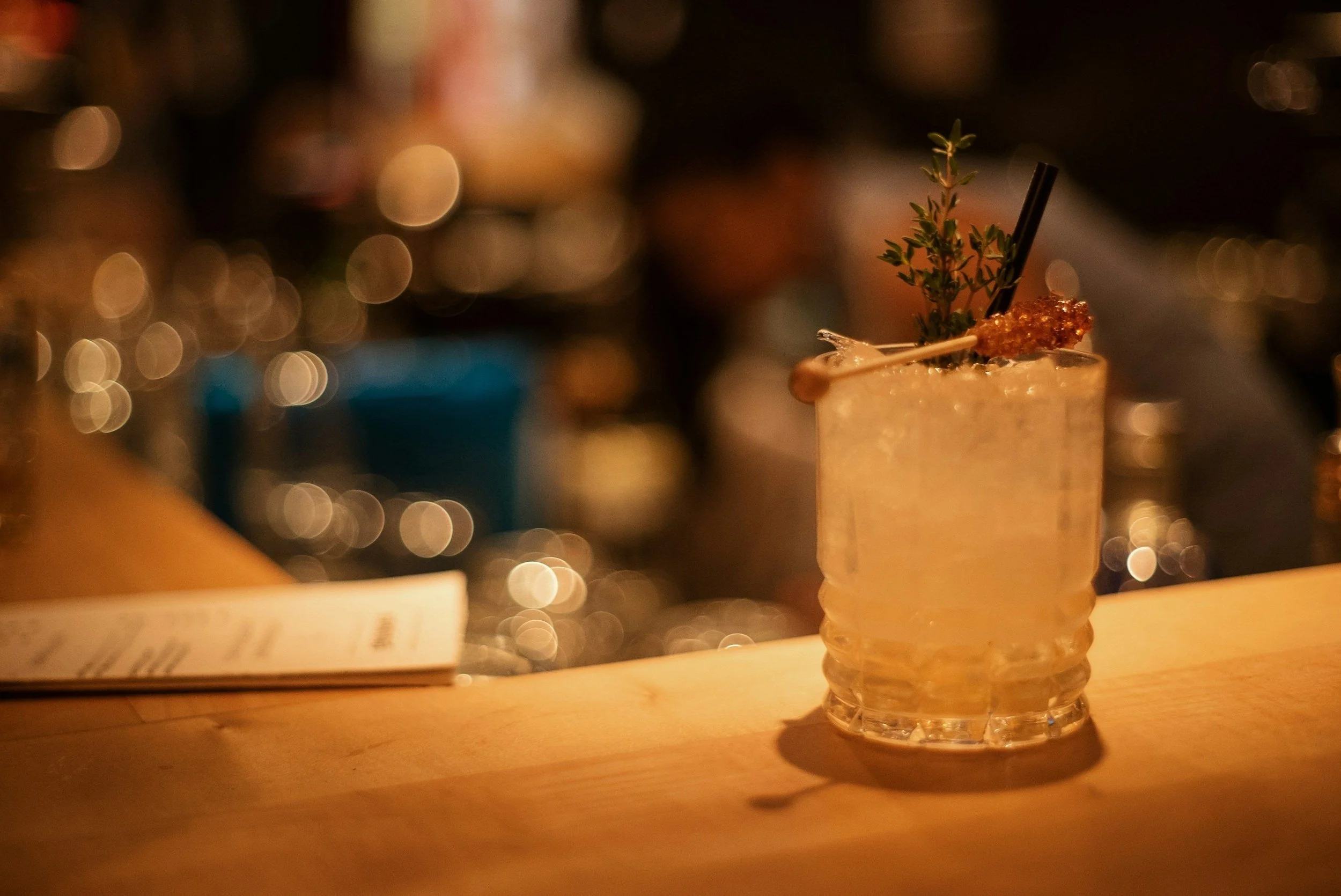 A cocktail with ice, garnished with herbs and a rock candy stirrer on a bar counter.