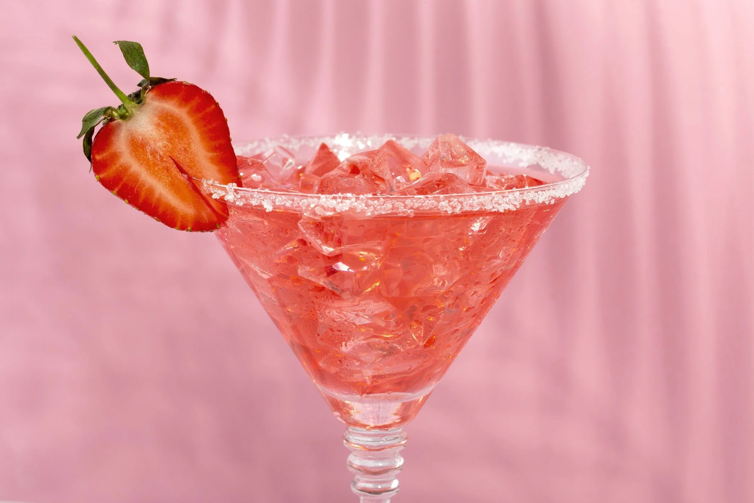 Pink cocktail in a martini glass with ice, garnished with a sliced strawberry on the rim against a pink background.