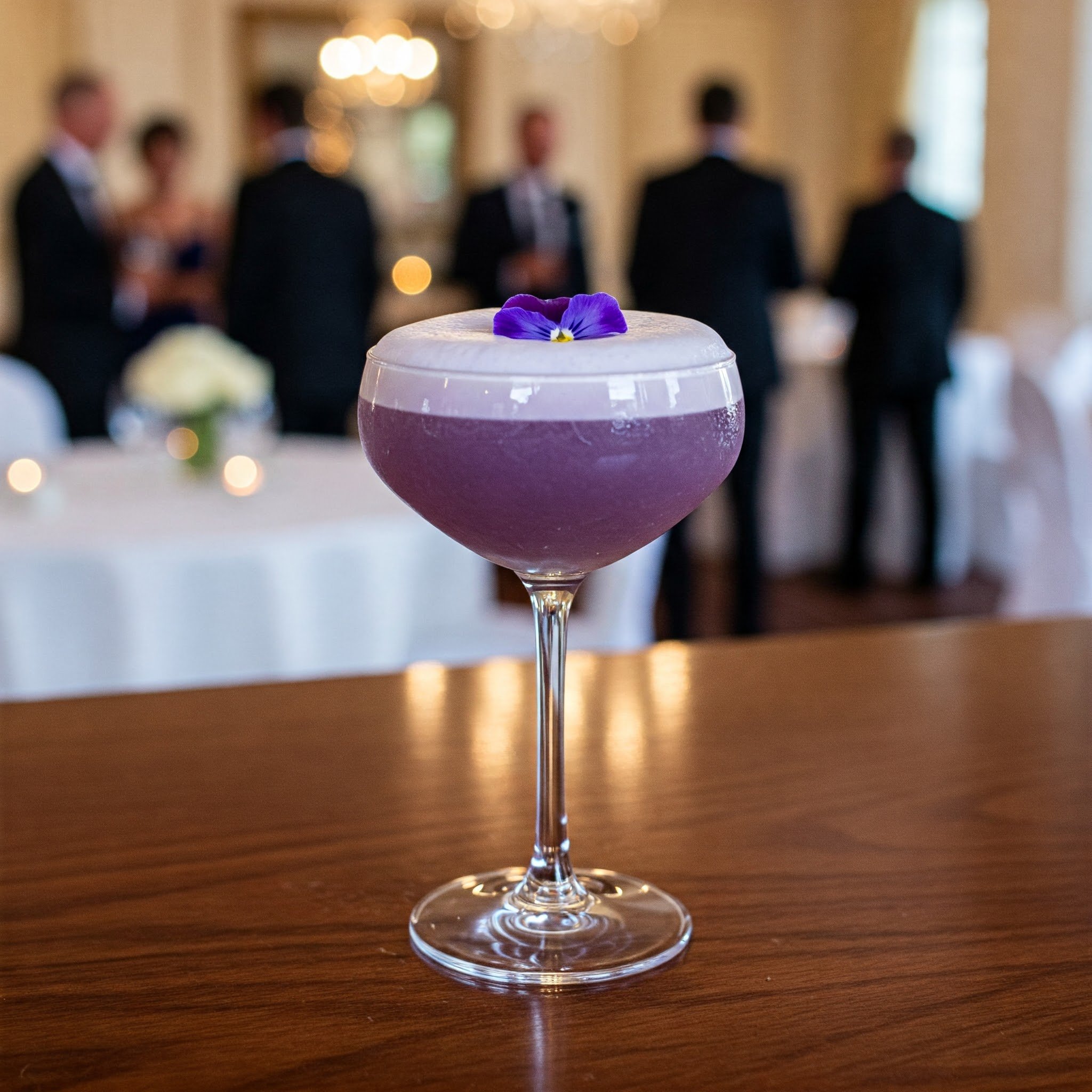 Purple cocktail in a coupe glass garnished with a purple flower, with a blurred background of people socializing at an event.
