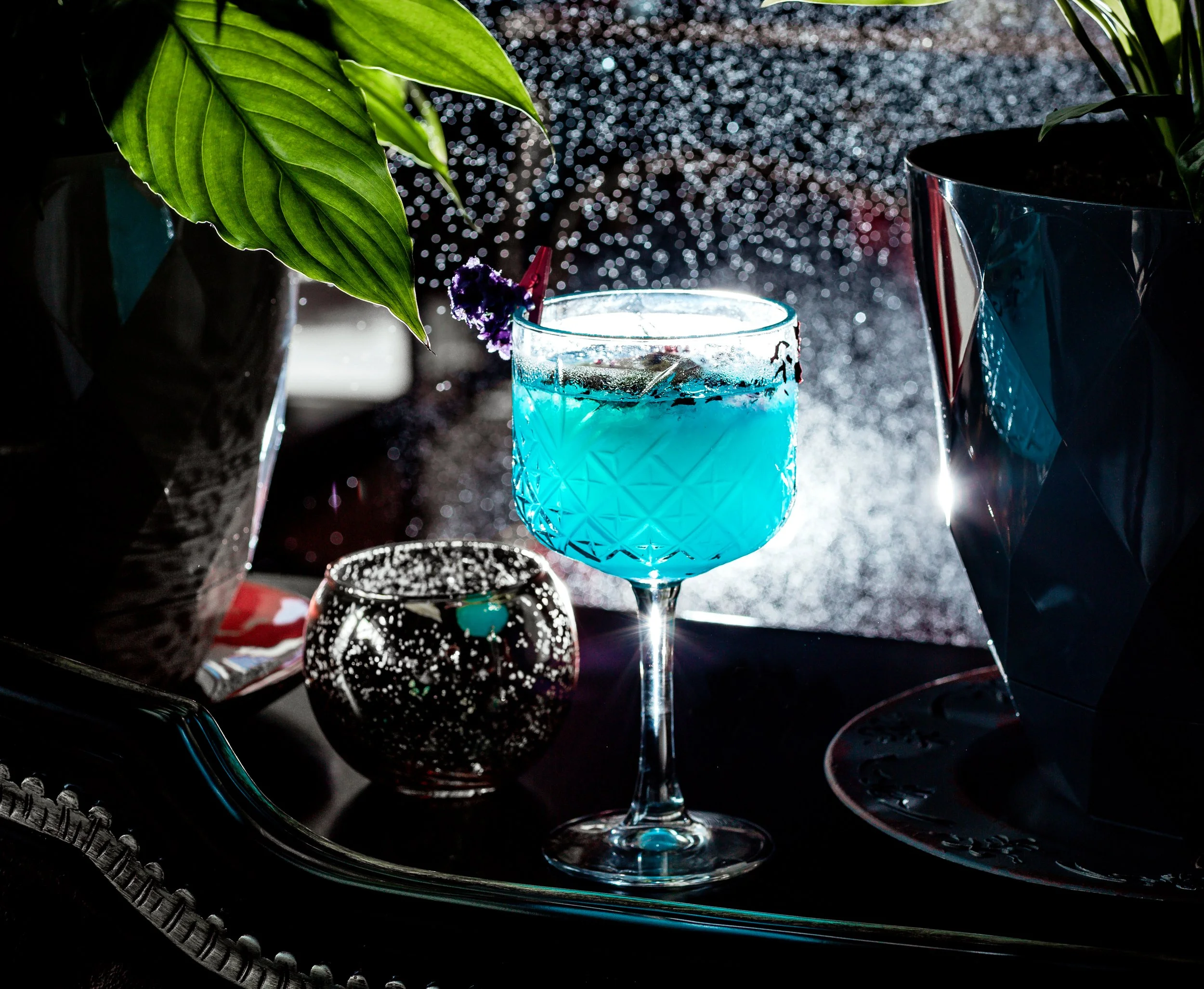 A blue cocktail in a decorative glass, surrounded by plants and a small patterned candle holder, with water droplets on the window in the background.