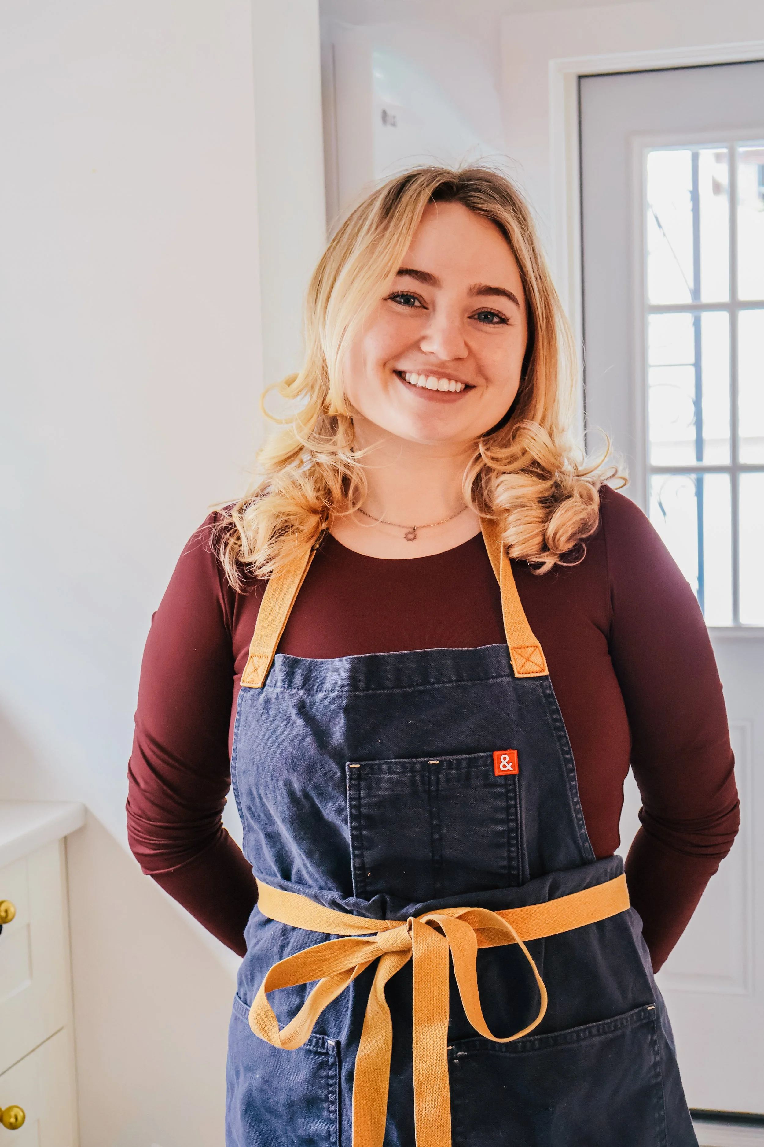 A woman with blonde hair smiling, wearing a maroon long-sleeved shirt, a denim apron with a red '&' logo, and a yellow apron tied around her waist, standing in a bright room near a window.