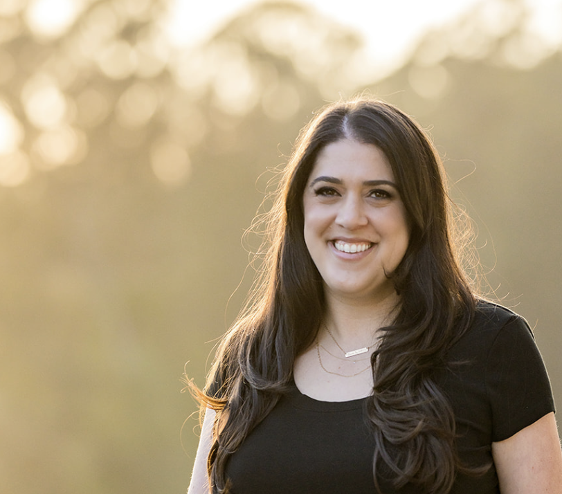 A smiling woman with long dark hair standing outdoors during sunset, wearing a black top.