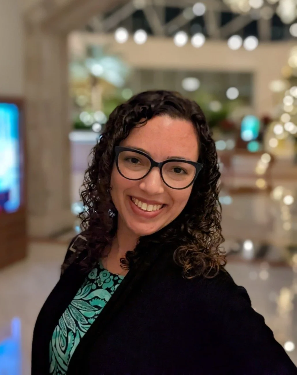 A woman with curly dark hair and glasses smiling at the camera, indoors with blurred lights in the background.