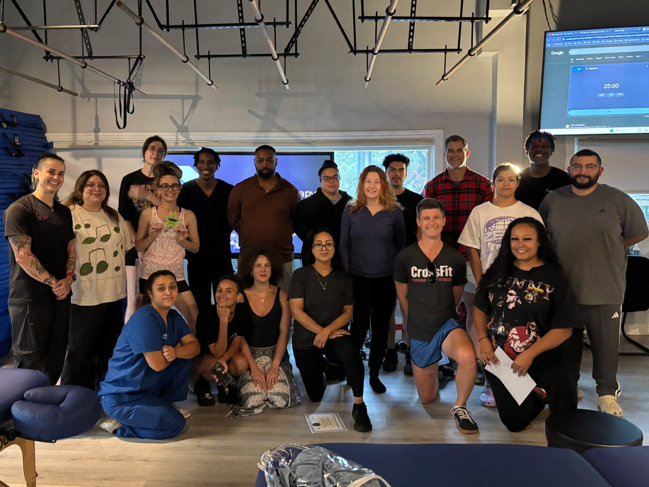 Group photo of smiling people in a fitness or community center, some kneeling and others standing, with exercise equipment and a screen in the background.