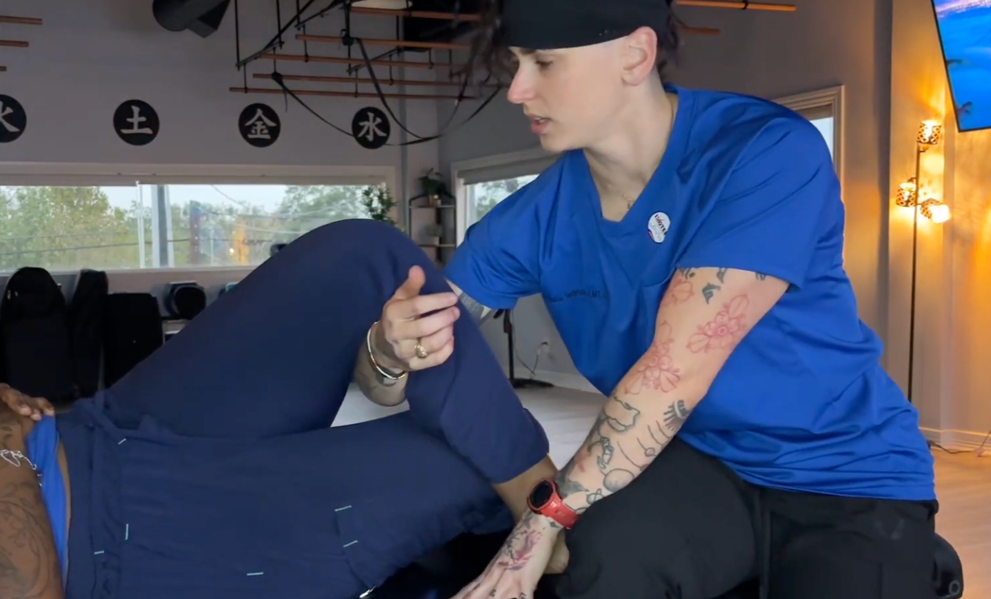 A physical therapy session showing a woman in a blue shirt assisting a person lying on their side, holding their arm for support, in a well-lit room with windows and Asian-themed decorations.