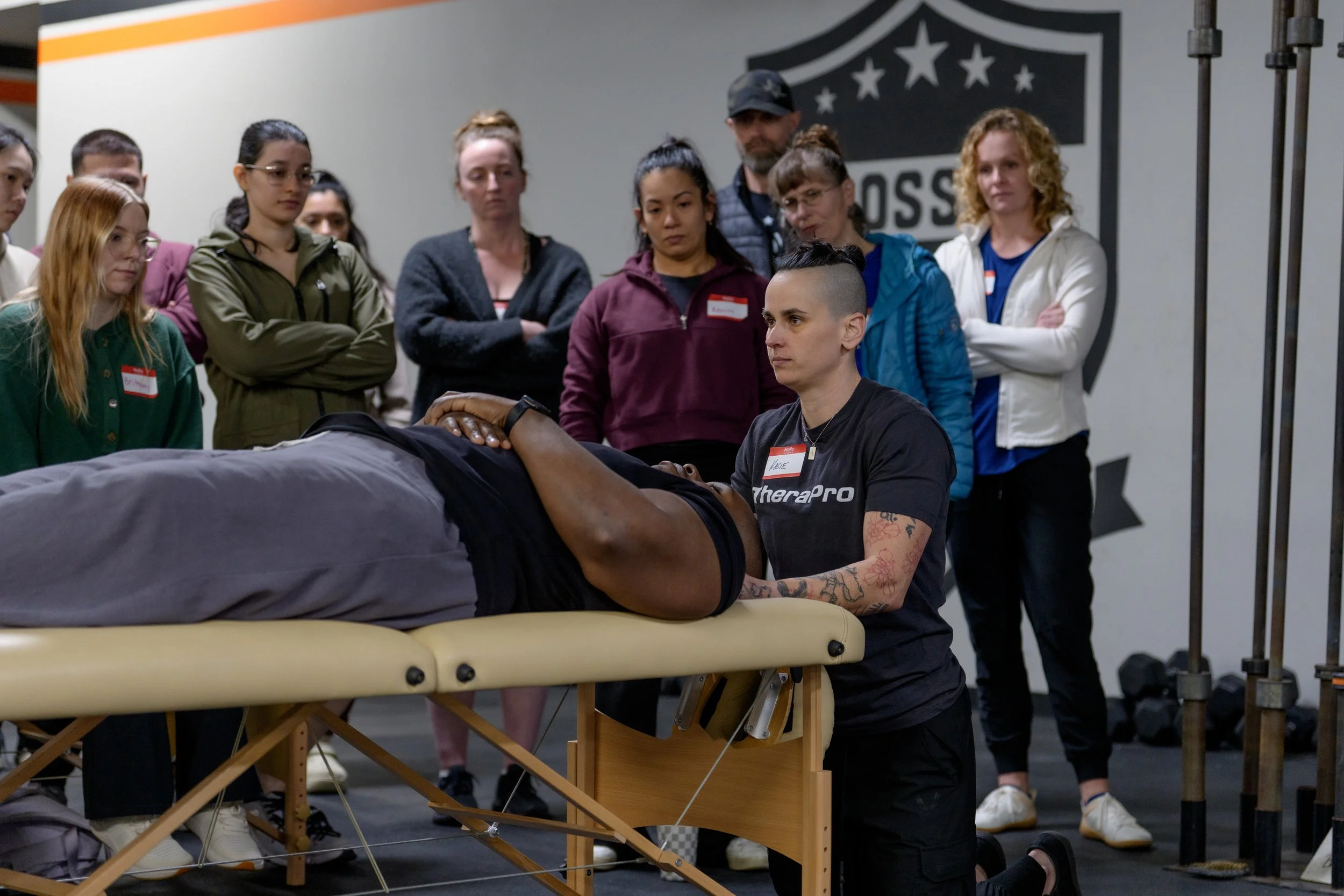 A group of women watching a person lying on a massage table during a training session, with one woman providing instruction.
