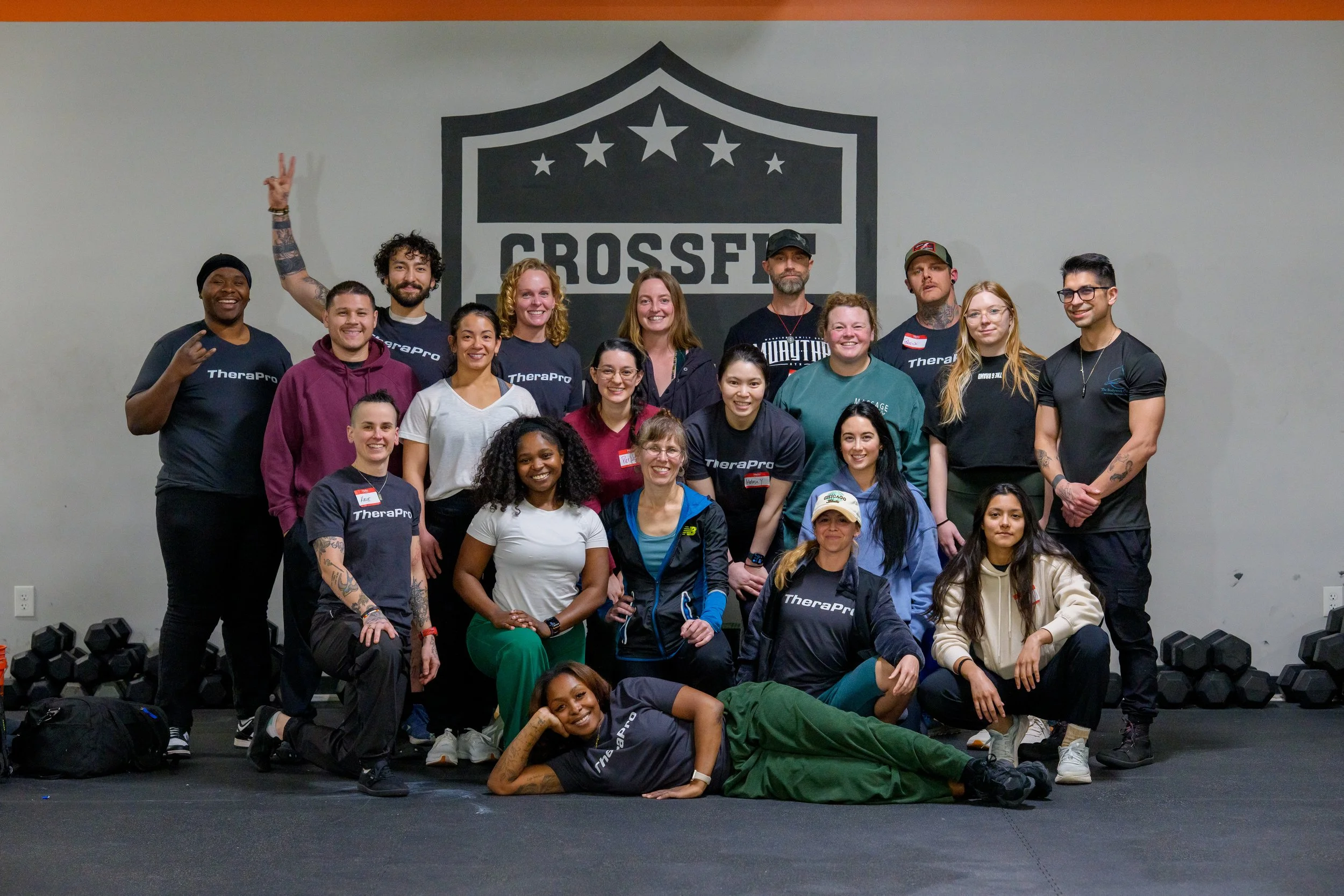 Group photo of smiling people in a fitness or community center, some kneeling and others standing, with exercise equipment and a screen in the background.