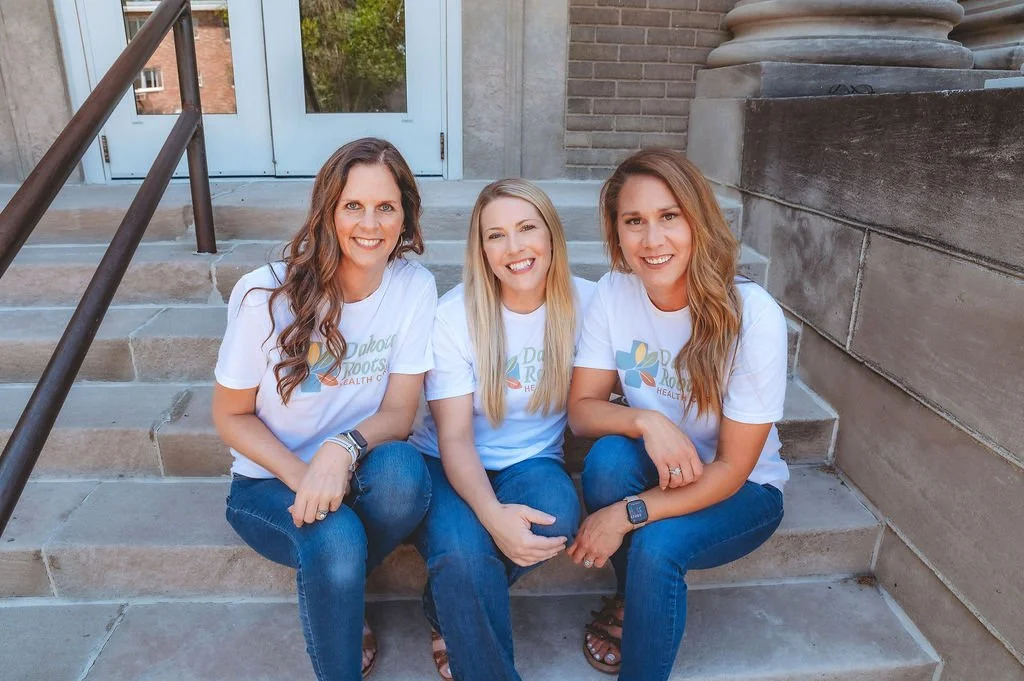 Three women sitting on steps outside a building, smiling at the camera, wearing matching white t-shirts with a logo and text.