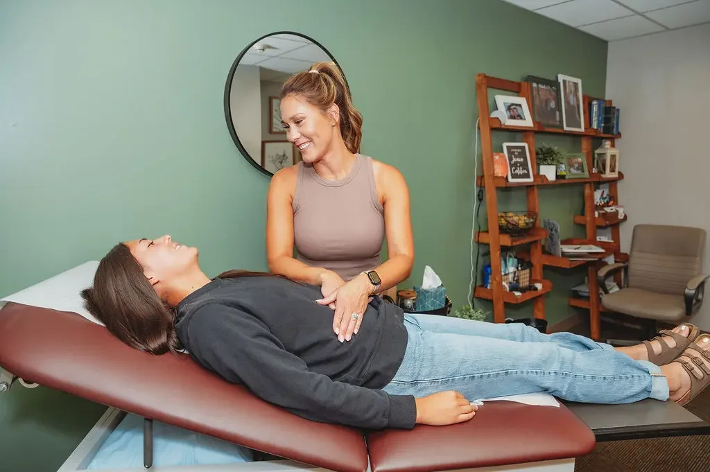 A woman lying on a medical examination table, smiling, while a healthcare provider stands beside her, also smiling, in a room with medical supplies and framed photos on a wooden shelf.
