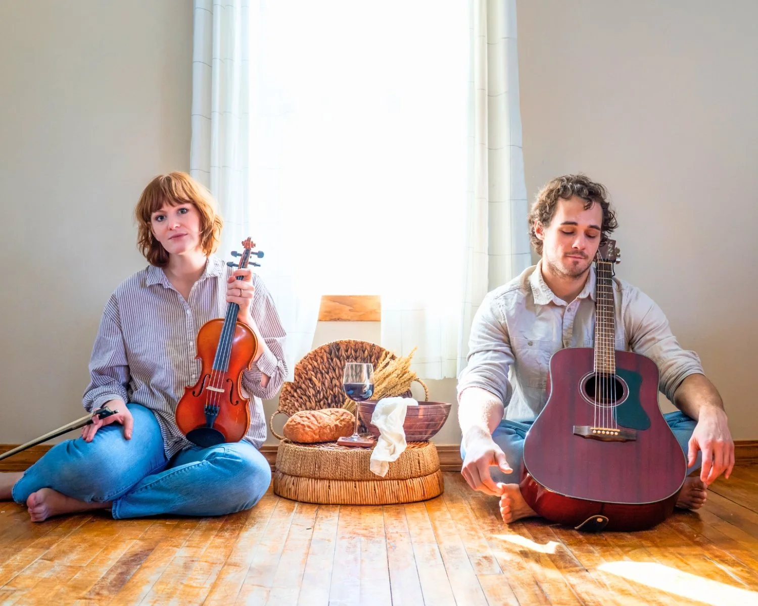Anna Leverett, a young woman holding a violin, and Damian Leverett, a young man with a guitar, sitting on a wood floor in a bright room with sunlight. A basket with a wine glass and loaf of bread on top is between them on the floor.