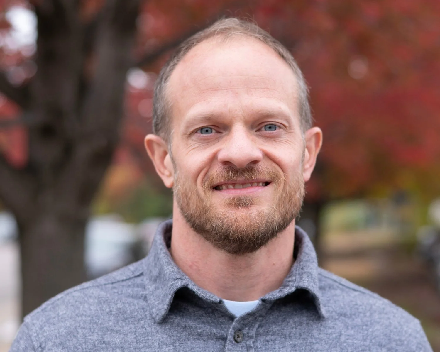 Portrait of Dave Omdal, a CityJoy staff member who mentors at-risk youth through Brazilian jiu-jitsu, smiling and standing in front of autumn trees with red leaves in the background.