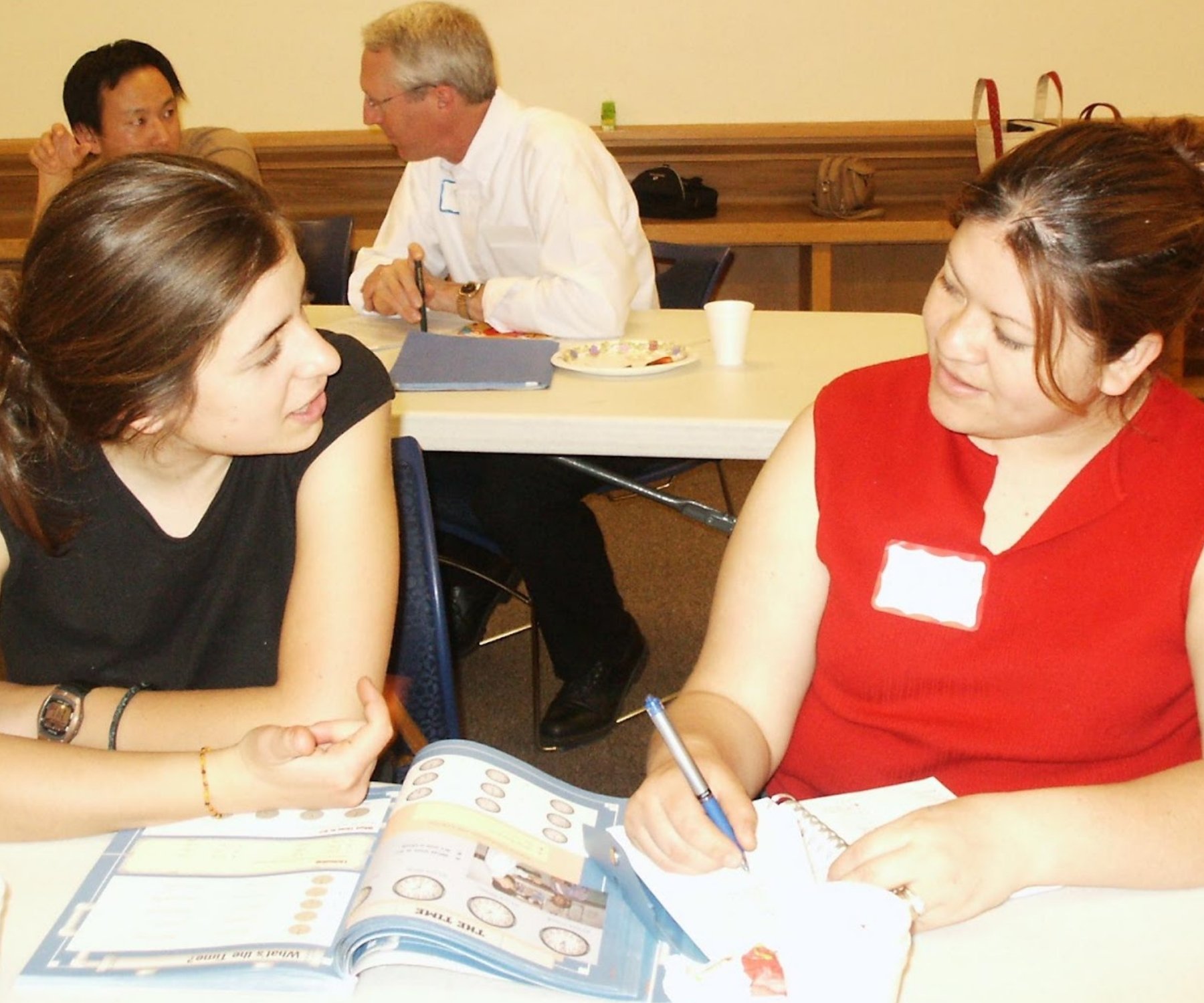 Two women having a conversation at a table with an ESL textbook and a notebook, while two people are in the background at another table.