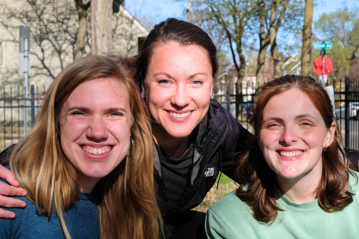 Three young women who help at Revive, a CityJoy ministry, smiling outdoors in front of a black fence with trees and houses in the background.