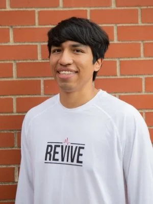 Portrait of Tony Dominguez, a program coordinator with Revive, a youth mentoring ministry connected to CityJoy in Minneapolis, smiling and standing in front of an orange brick wall.