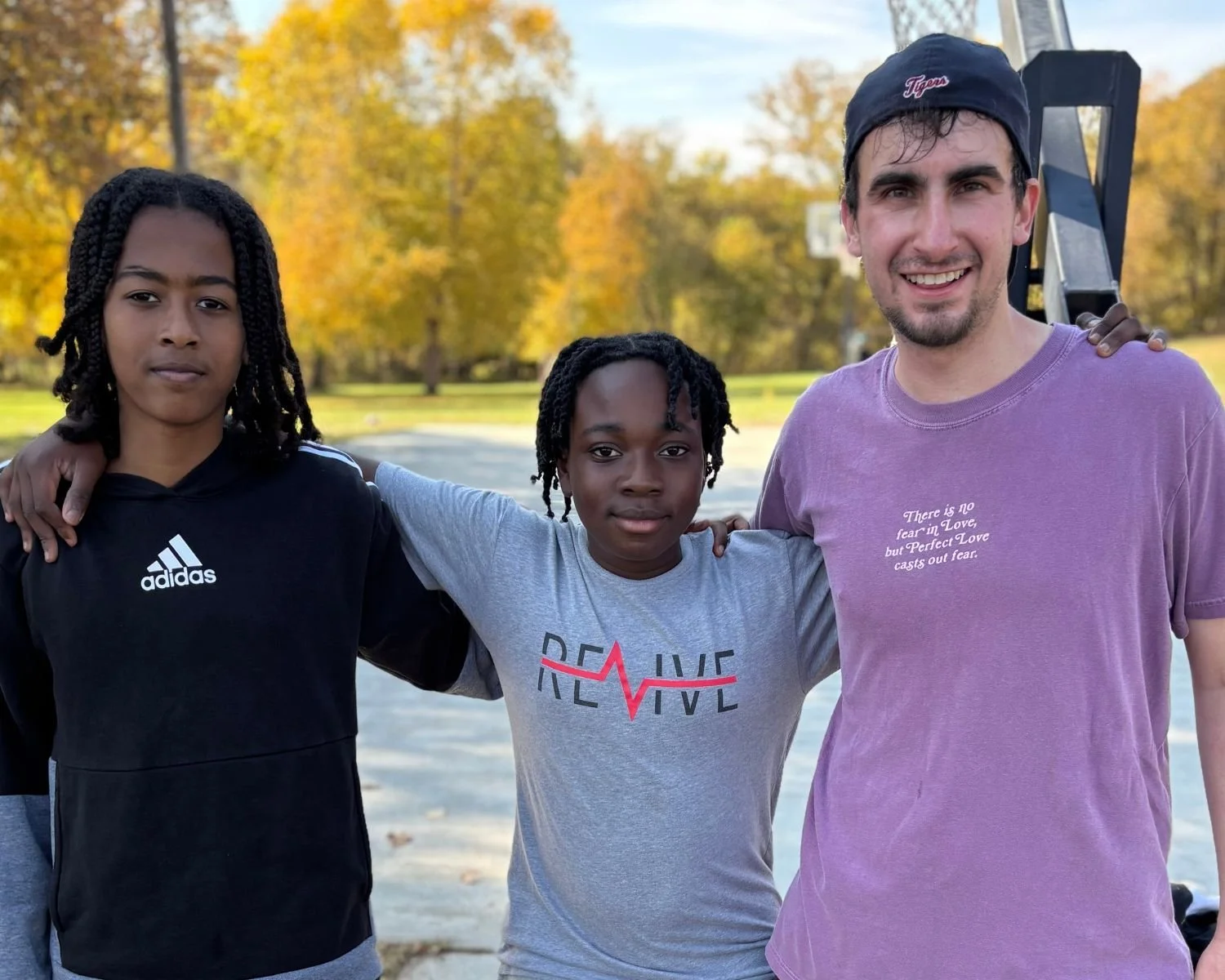 Three people standing together outdoors on a basketball court with fall foliage in the background and their arms around each others shoulders. There is a teen boy on the left and middle. On the right is a man who is a youth mentor for Revive.