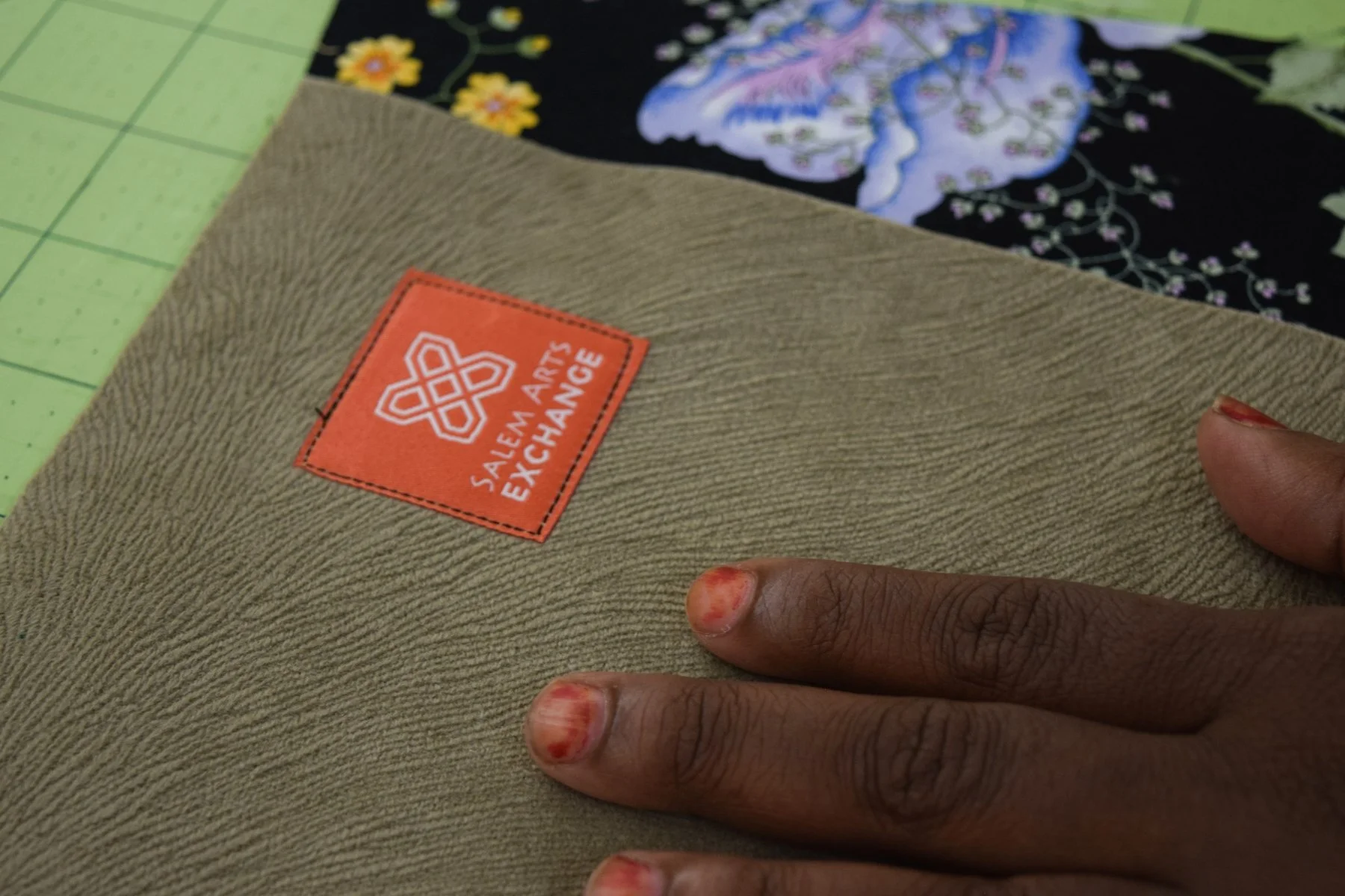A person's hand with red finger nail polish resting on a textured beige surface with an orange label that reads 'Salem Arts Exchange' and features a white geometric logo. Part of a colorful fabric with floral and abstract patterns is visible in the b