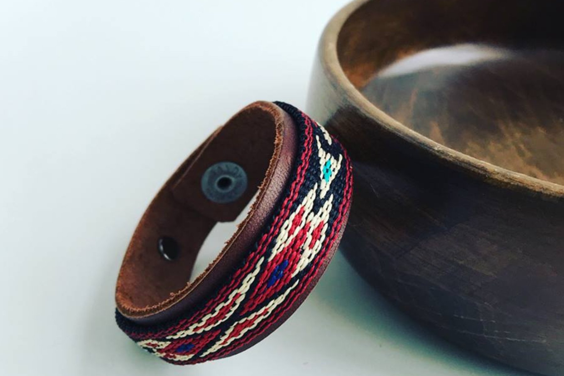 A leather bracelet with colorful embroidery hanging on the edge of a dark wooden bowl, placed on a white surface.
