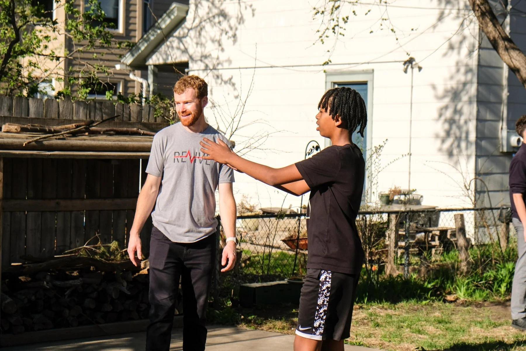 Two young men standing outside, one touching the other's chest, engaged in conversation, with a backyard and house in the background.