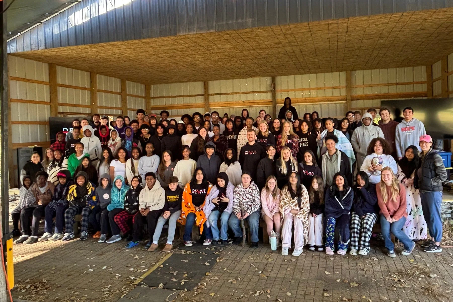 A large group of diverse young people gathers indoors for a group photo, some sitting in the front and others standing behind, with wood-paneled walls and a ceiling in the background.