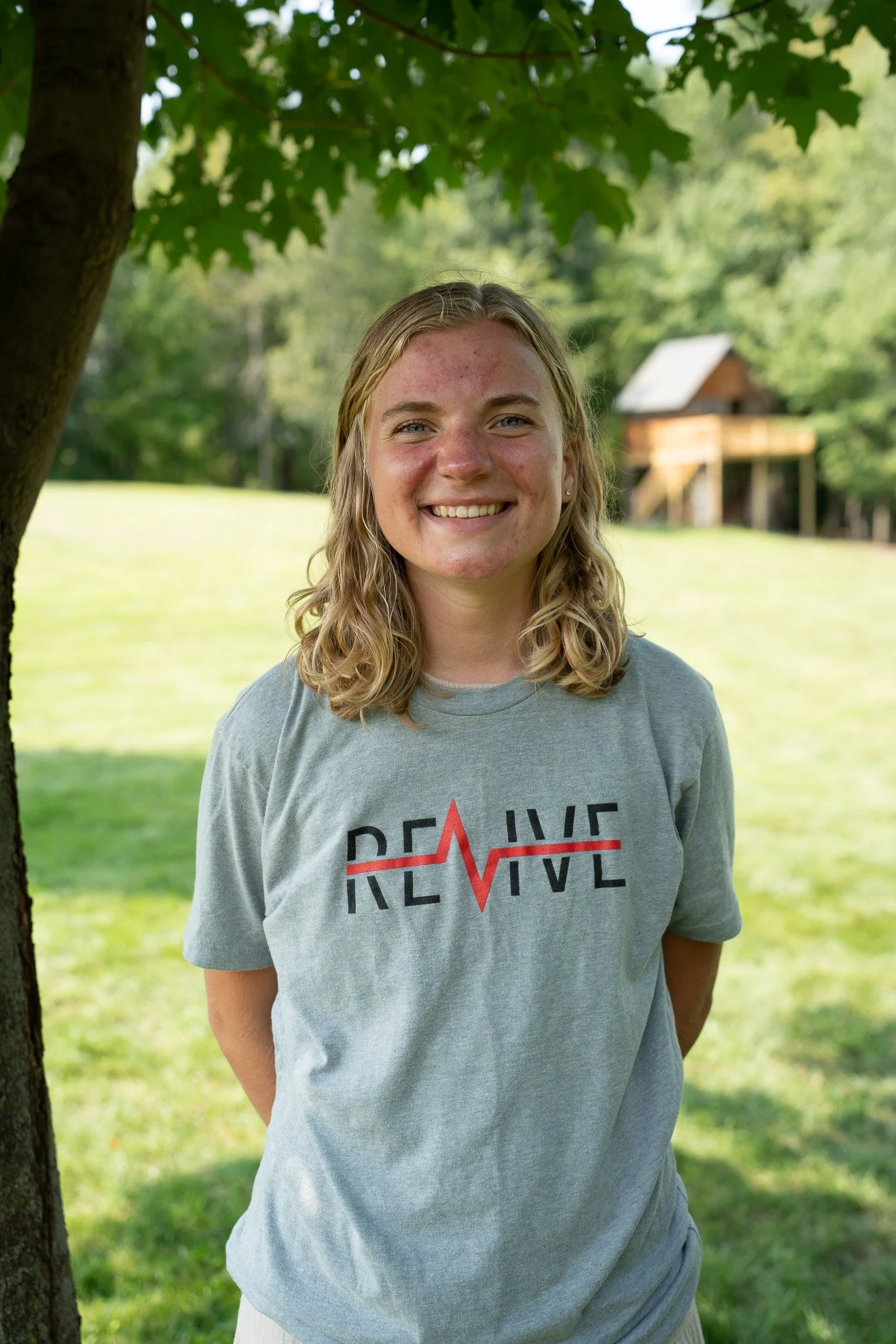 Portrait of Sam Erickson, a CityJoy staff member who works with Revive and mentors youth, smiling outdoors and standing under to a tree with a grass field in the background.