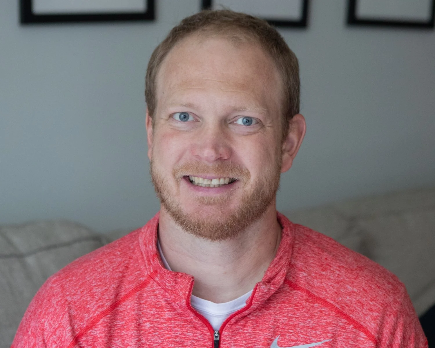 Aaron Robertson, a CityJoy minister with blue eyes and a beard, smiling and wearing a red athletic shirt, sitting on a gray couch in a room with black picture frames on the wall.