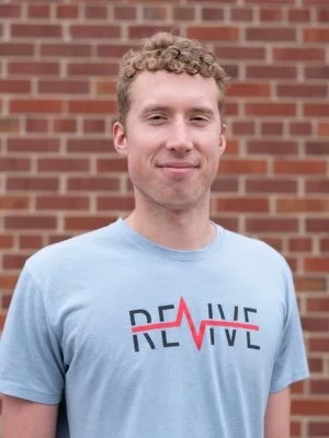 Portrait of Sam Erickson, a program coordinator with Revive, a youth mentoring ministry connected to CityJoy in Minneapolis, smiling and standing in front of an orange brick wall.
