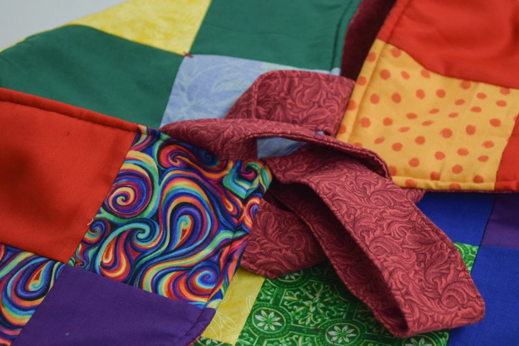 Close-up of a colorful quilted pot holder at Salem Arts Exchange with various bright patterned fabric squares, including swirls, polka dots, and solid colors, with a tie on top.
