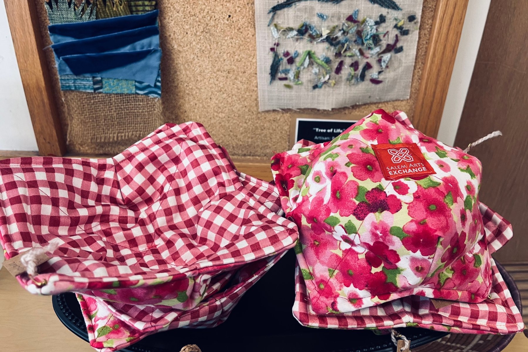 Two fabric pot warmers, one with a red and white checked pattern and the other with a pink floral pattern labeled 'Salem Arts Exchange,' on a black tray. In the background, there is a bulletin board with various textile art pieces.