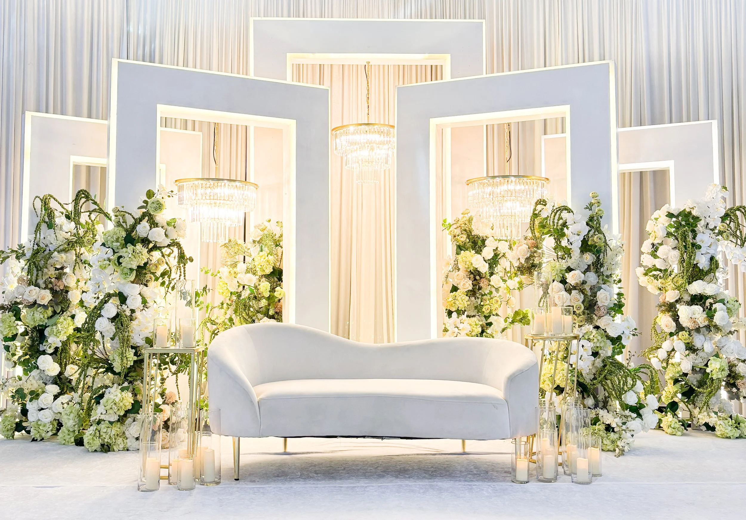 A decorated stage with a white sofa flanked by floral arrangements, a backdrop with red and cream curtains, and elegant lighting fixtures.