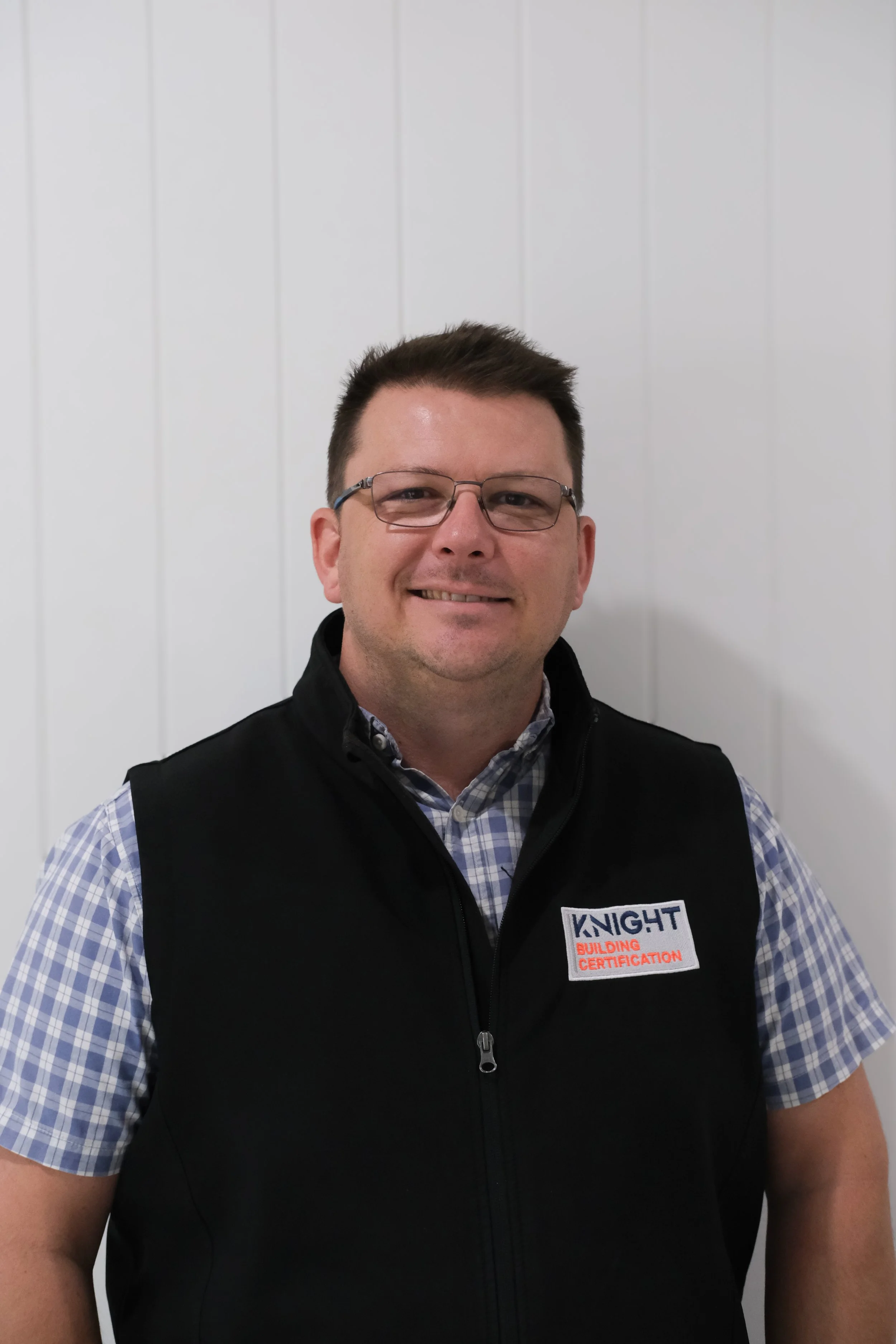 A man with short brown hair and glasses smiling, wearing a black vest over a blue and white checkered shirt, standing in front of a plain white wall. The vest has a label reading 'Knight Building Certification'.