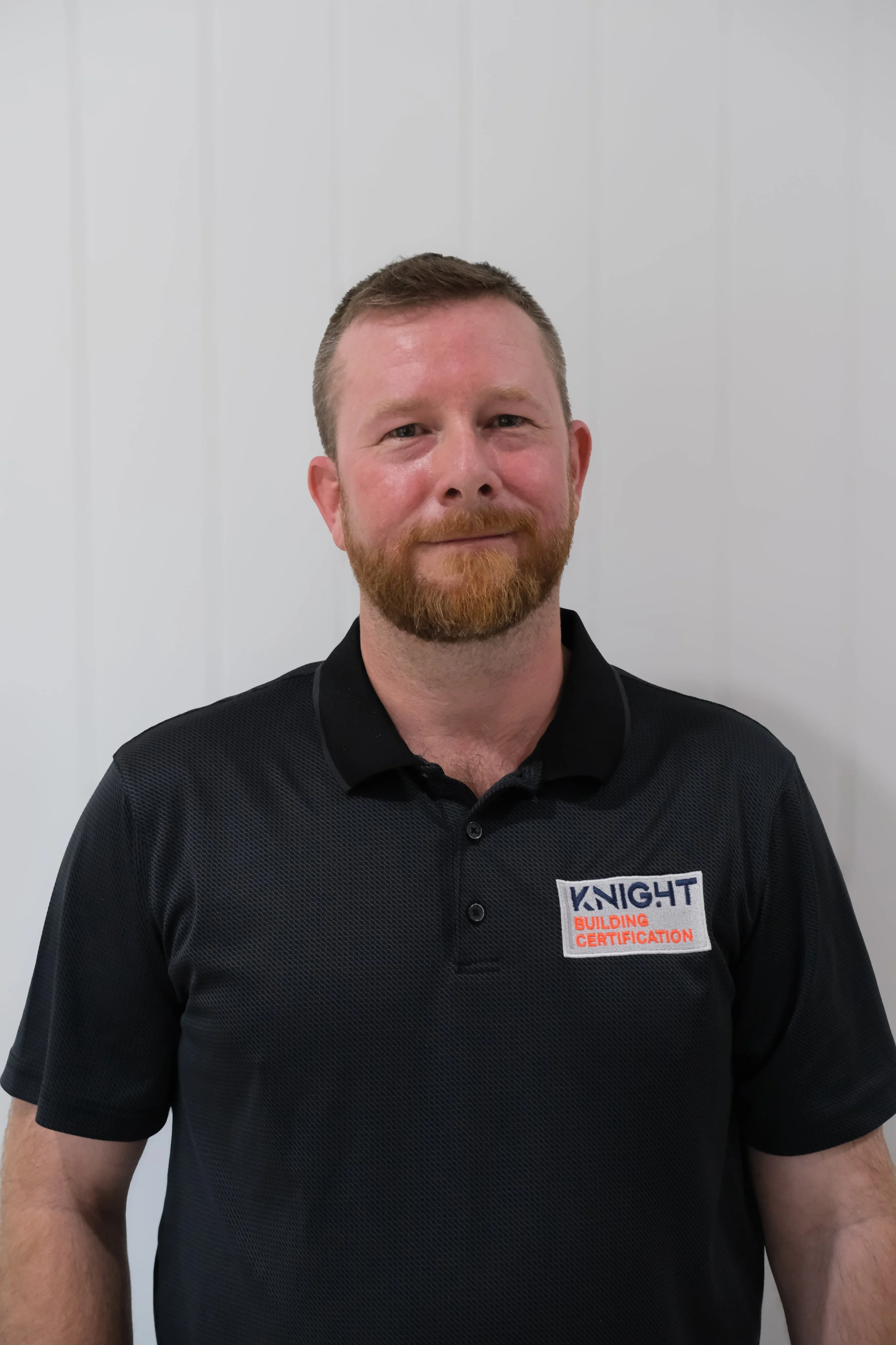 A man with short hair and a beard, wearing a black polo shirt with a patch that says "KNIGHT BUILDING CERTIFICATION," standing against a plain white wall.