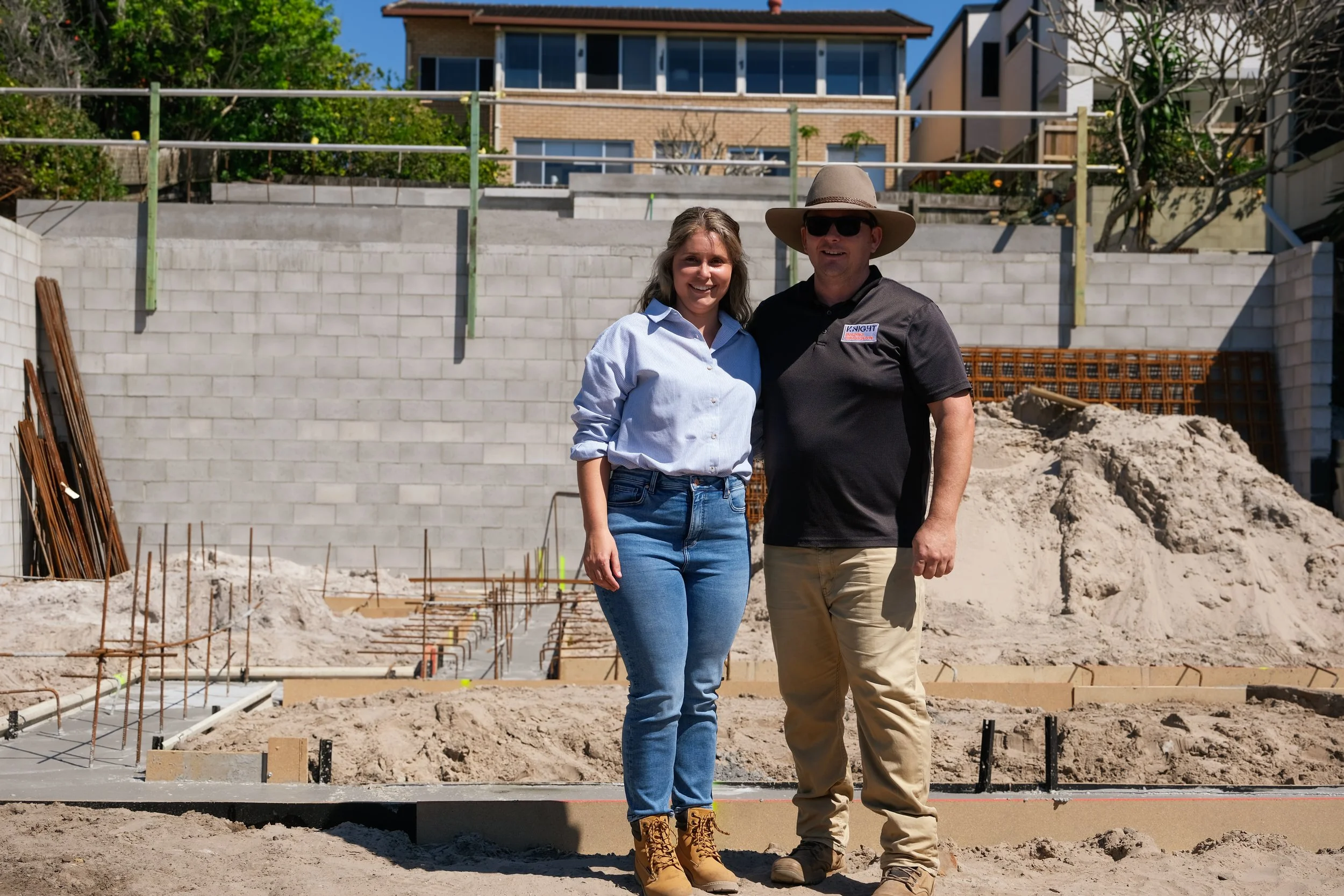 A man and a woman standing together at a construction site with a partially built foundation and a concrete block wall behind them. The woman is smiling and wearing a light blue button-up shirt and jeans, while the man is wearing a black polo shirt, khaki pants, sunglasses, and a wide-brimmed hat. Residential buildings and clear blue sky are visible in the background.