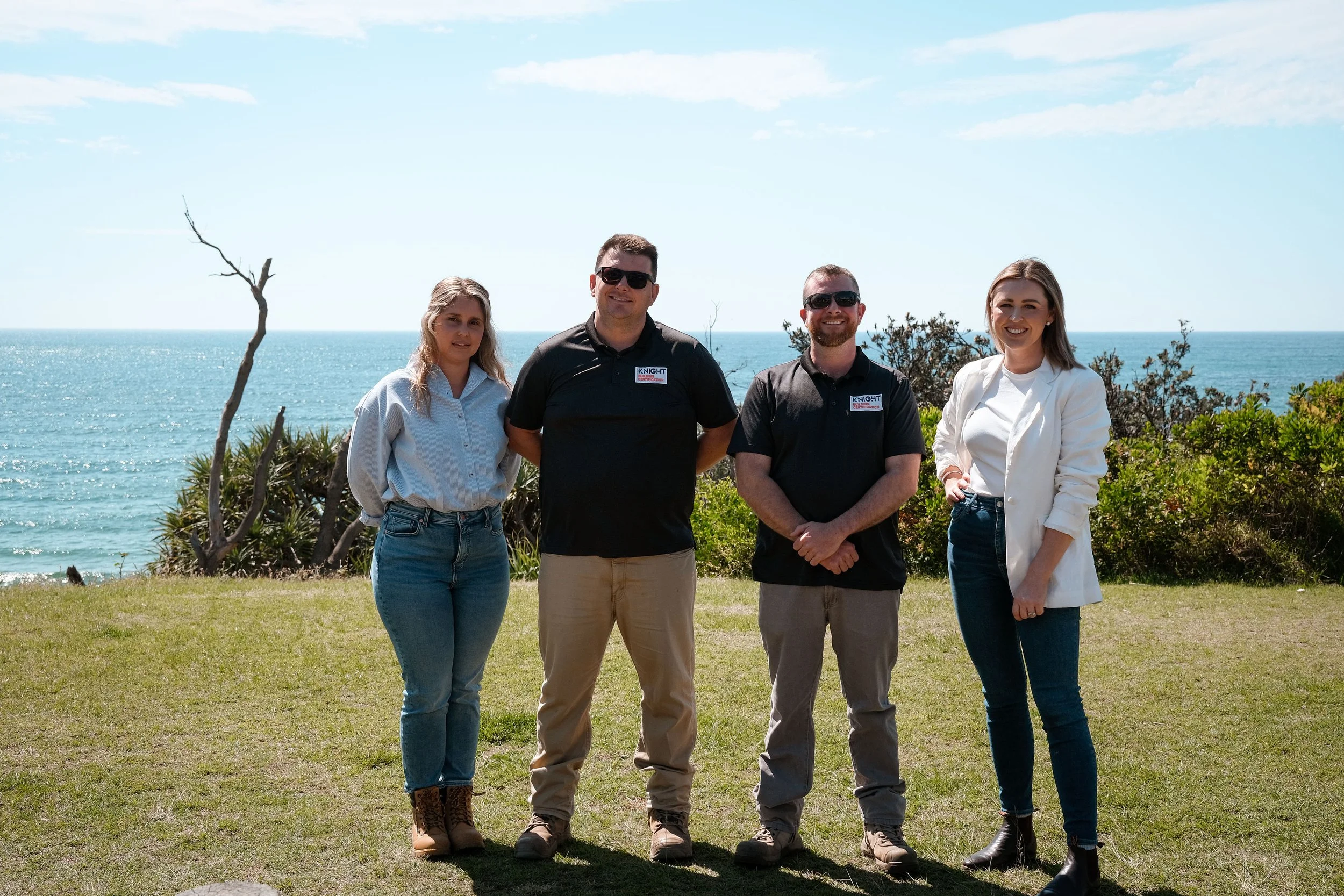 Four people standing outdoors near the ocean, dressed casually, smiling at the camera, with a blue sky and some greenery in the background.