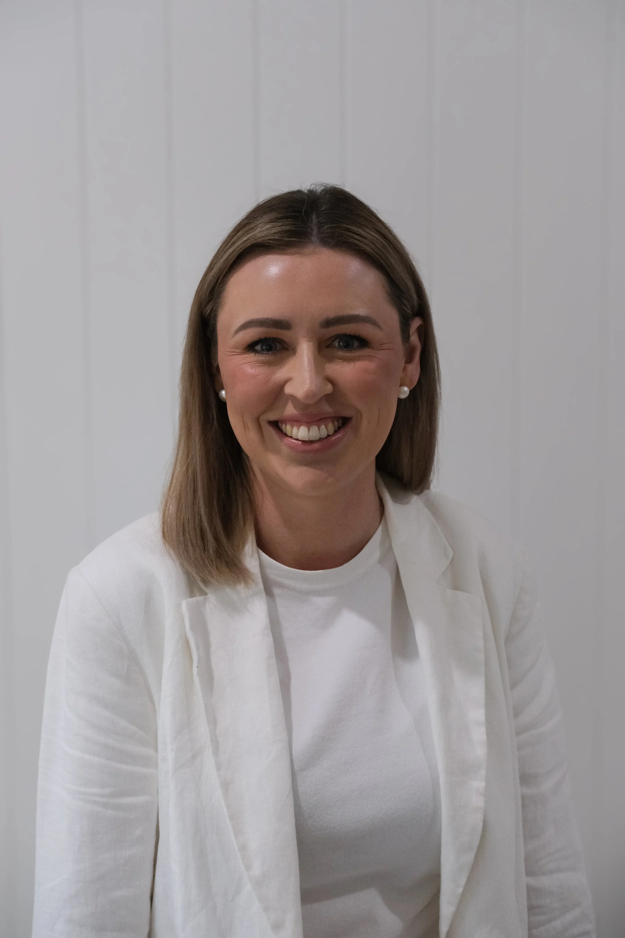 A woman with blonde hair wearing pearl earrings, a white blazer, and a white top, smiling in front of a plain, light-colored background.