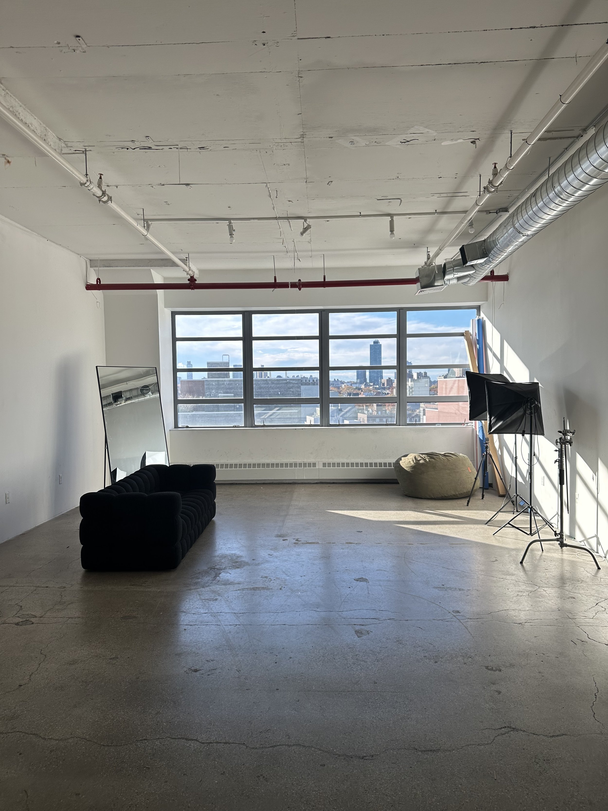 Interior of a photo studio with a large window, a black sofa, a mirror, photography lighting equipment, and a bean bag