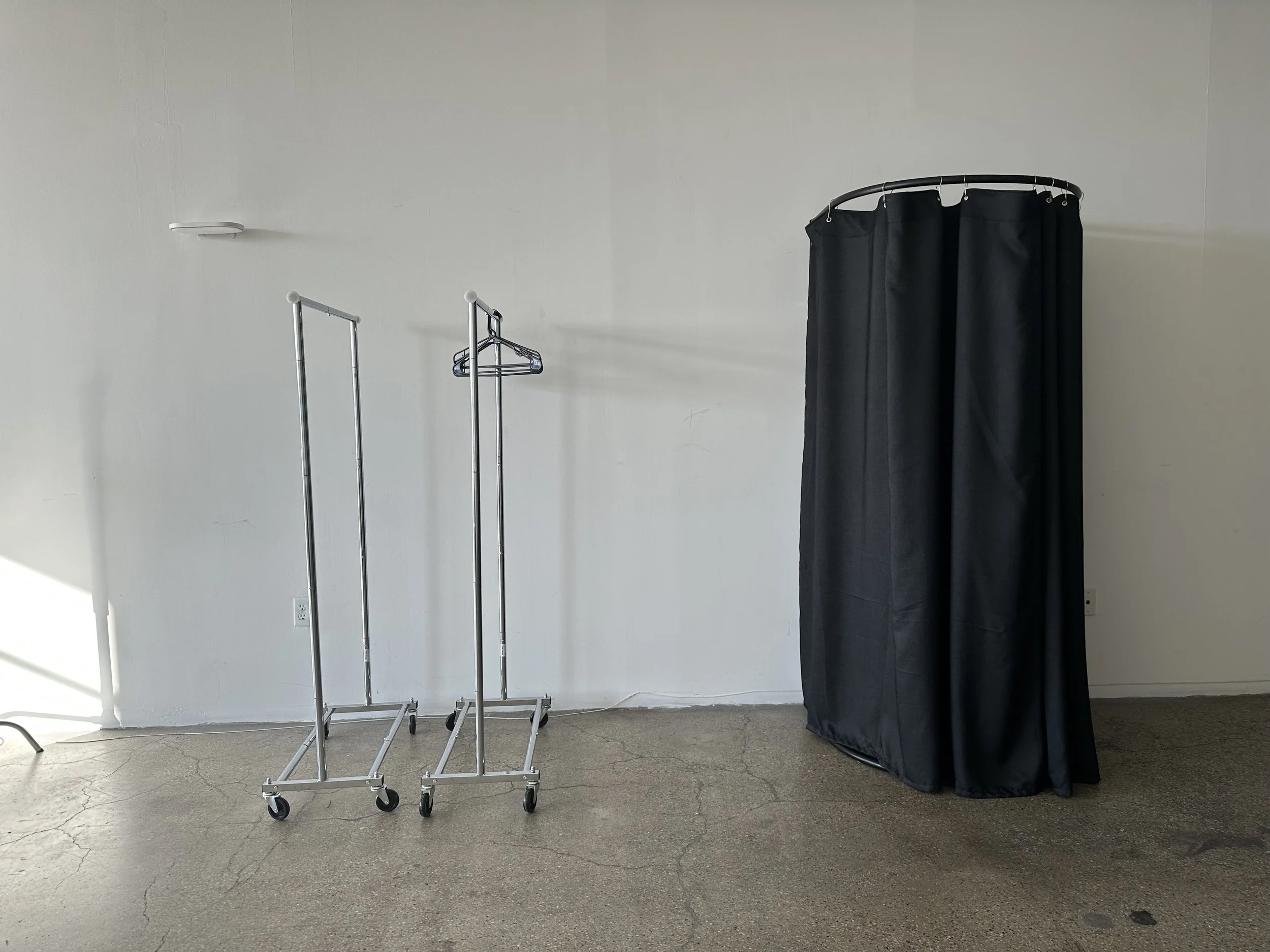 Two metal rolling garment racks next to a black fabric changing room curtain on a wall in a room with plain white walls and cracked concrete floor.