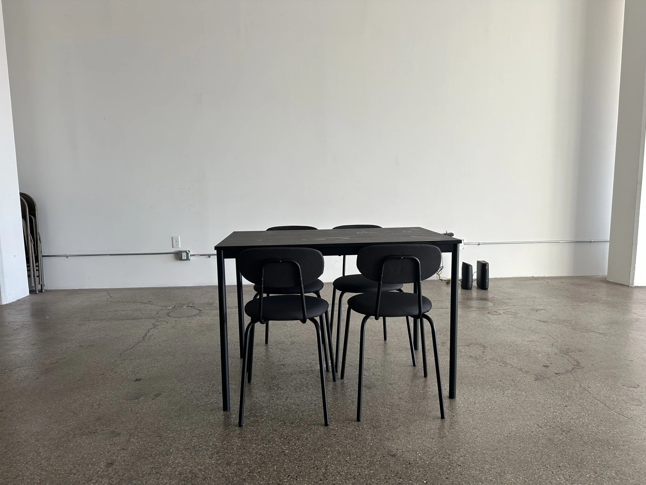 A black rectangular table with four black chairs in a minimalist industrial room with a plain white wall, concrete floor, and electrical outlets.