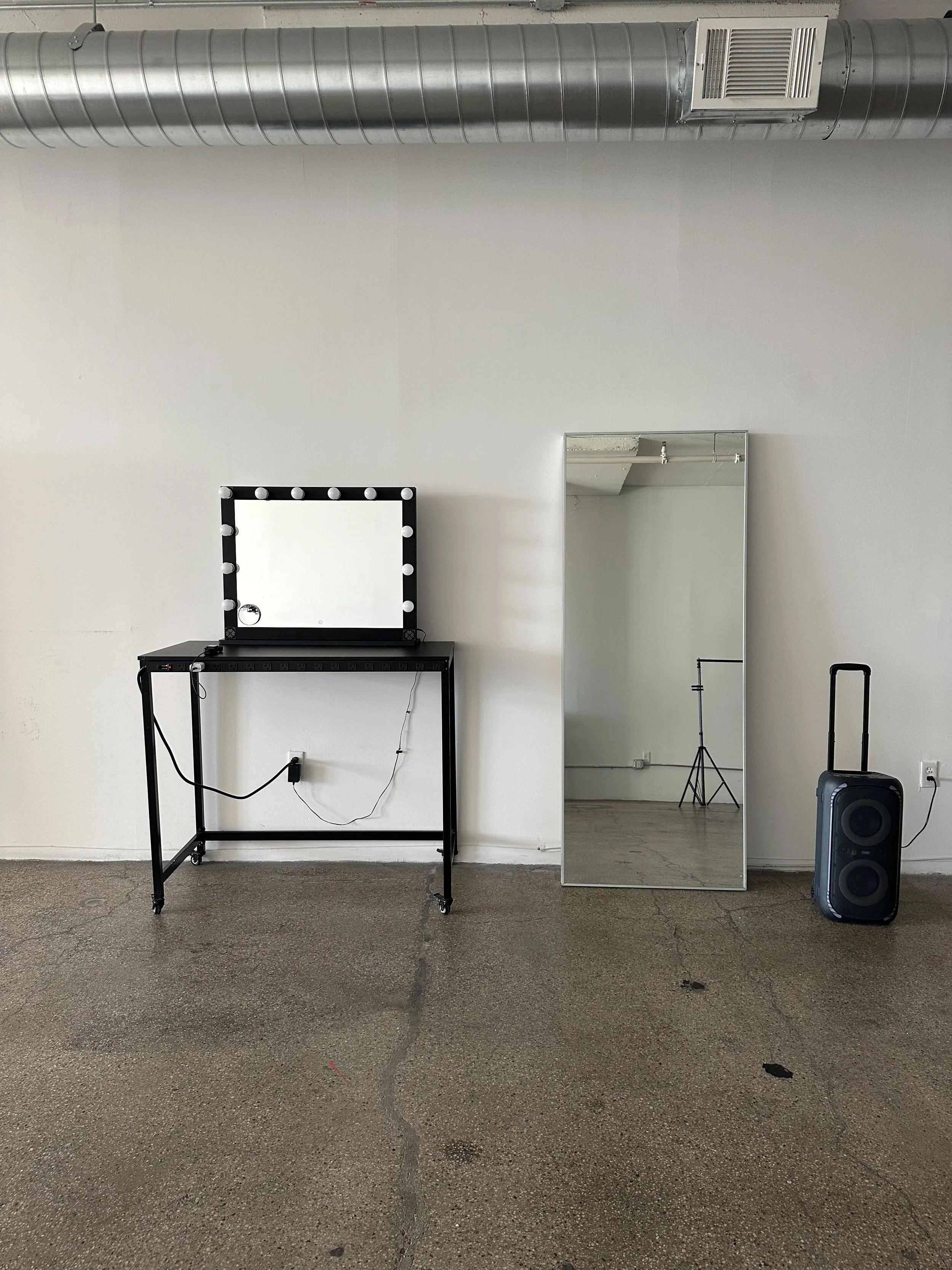 A makeup vanity station with a mirror surrounded by lights on a black table, a tall mirror to the right, and a portable speaker on the floor to the right. There is also a tripod visible in the reflection of the tall mirror. The room has a bare wall, 