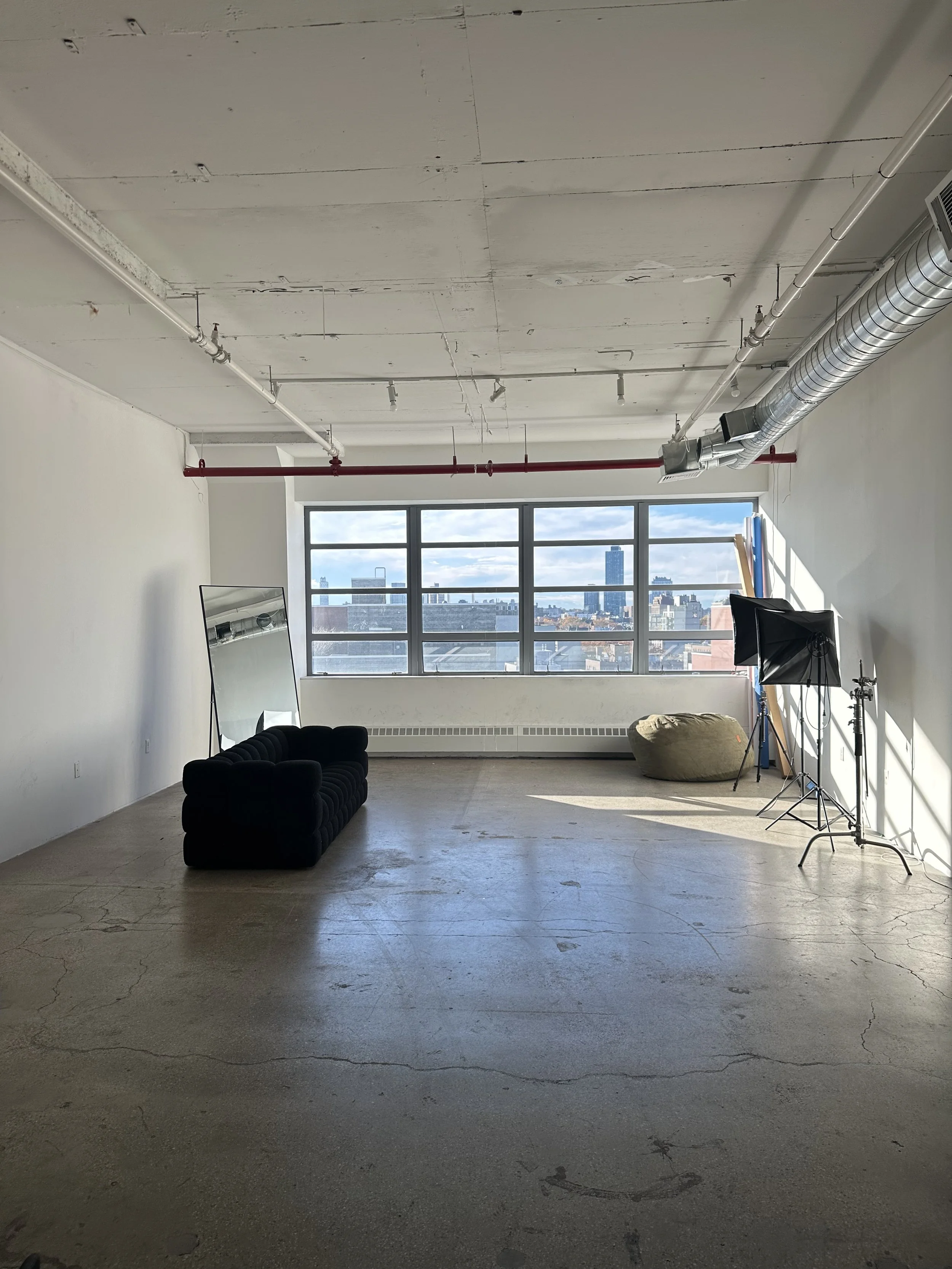 Empty room with large windows showing city skyline, black sofa, mirror, lighting equipment, and a bean bag.