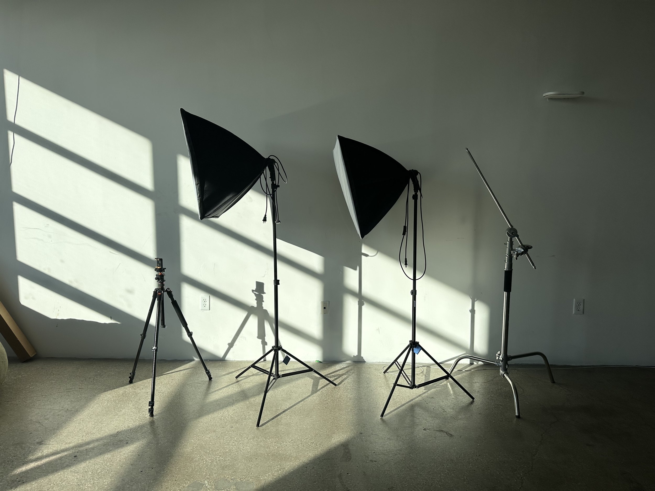 Photography studio setup with two large softbox lights, a camera on a tripod, and a light stand, illuminated by sunlight coming through a window.