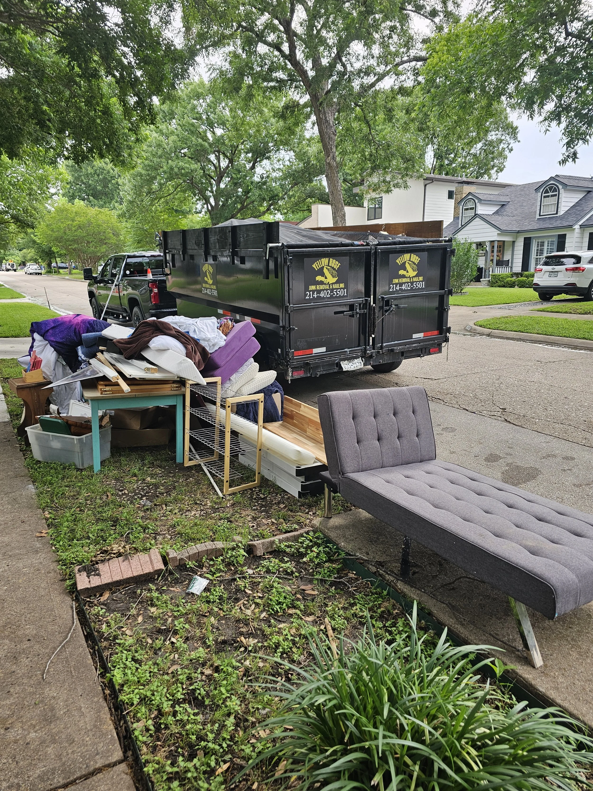 Collection of discarded furniture and household items on the sidewalk, including a gray chaise lounge, a wooden bookcase, a metal rack, a plastic container, and pillows, with a black dumpster in the background, surrounded by trees and residential hou