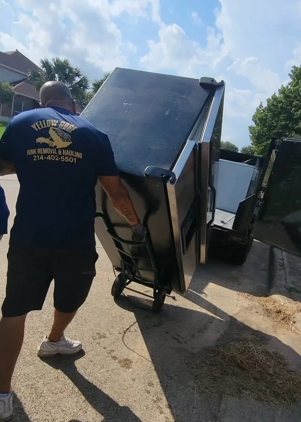 A worker with yellow rose logo is moving a large black refrigerator removing from house in Dallas.