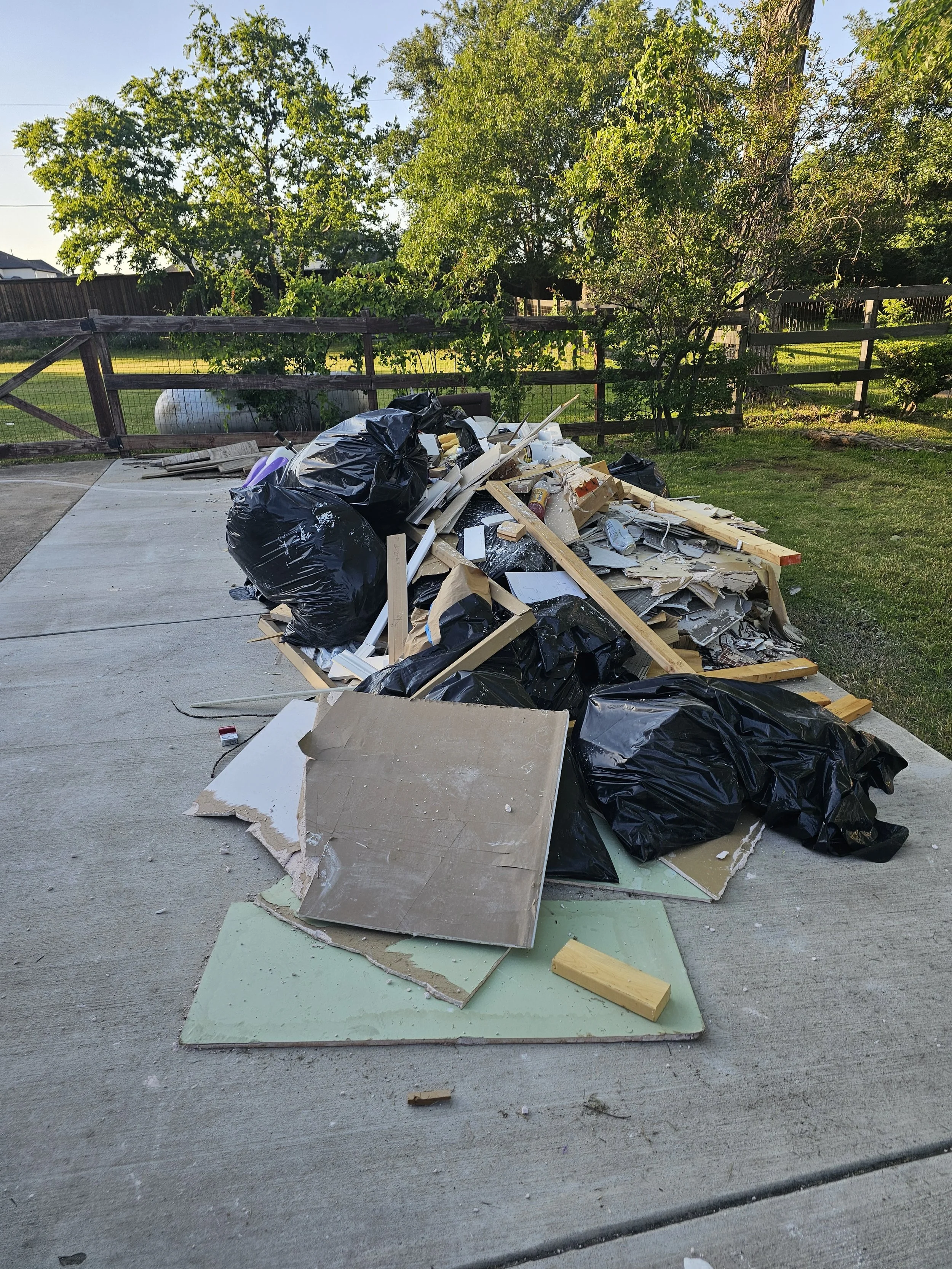 Piles of construction debris and trash including wood, drywall, cardboard, and black plastic bags on a concrete driveway, with a grassy yard and wooden fence in the background.
