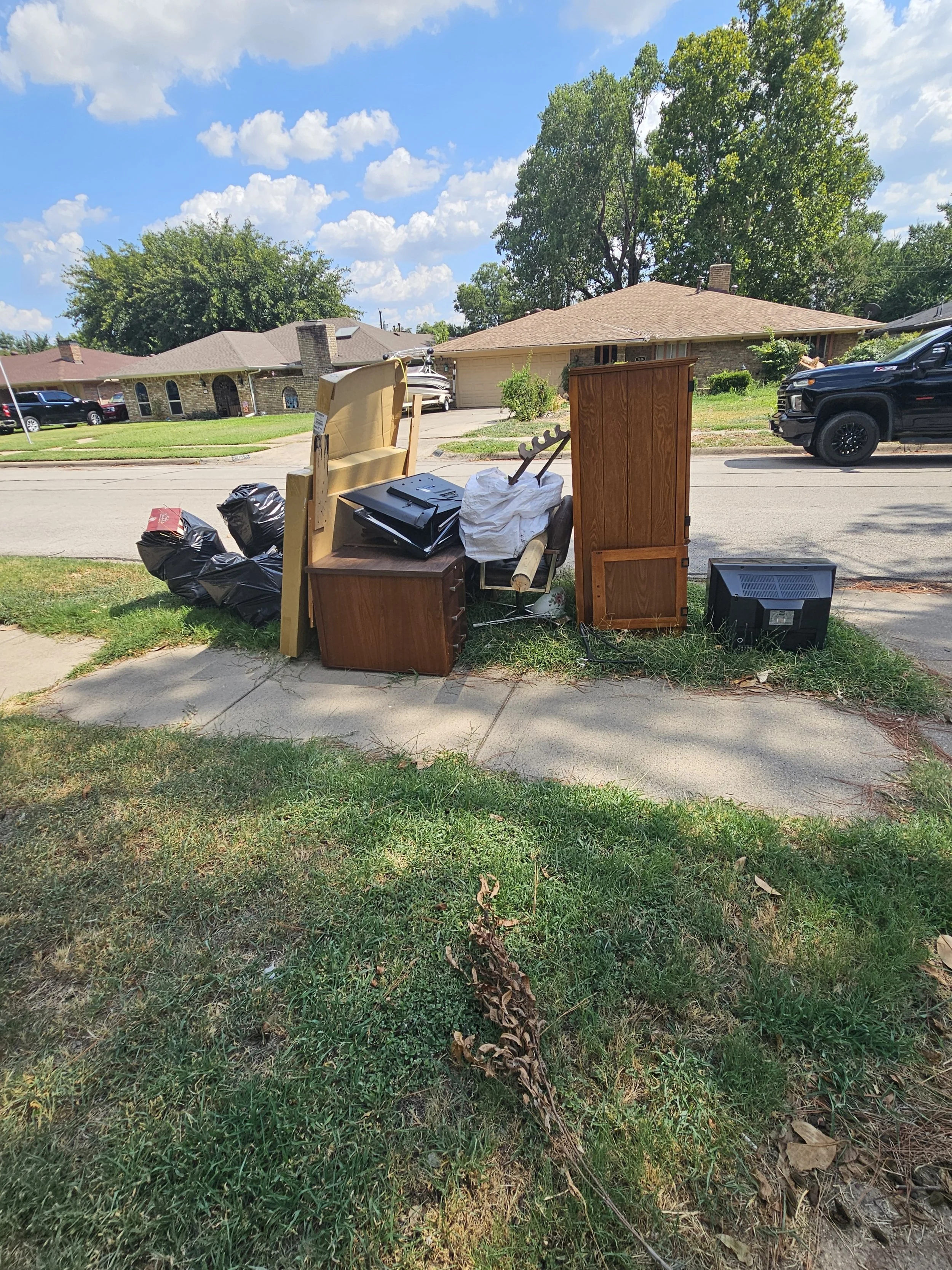 Collection of discarded furniture and household items on the sidewalk, including a headboard, a small dresser, a plastic bag with trash, a wooden cabinet, and an old television
