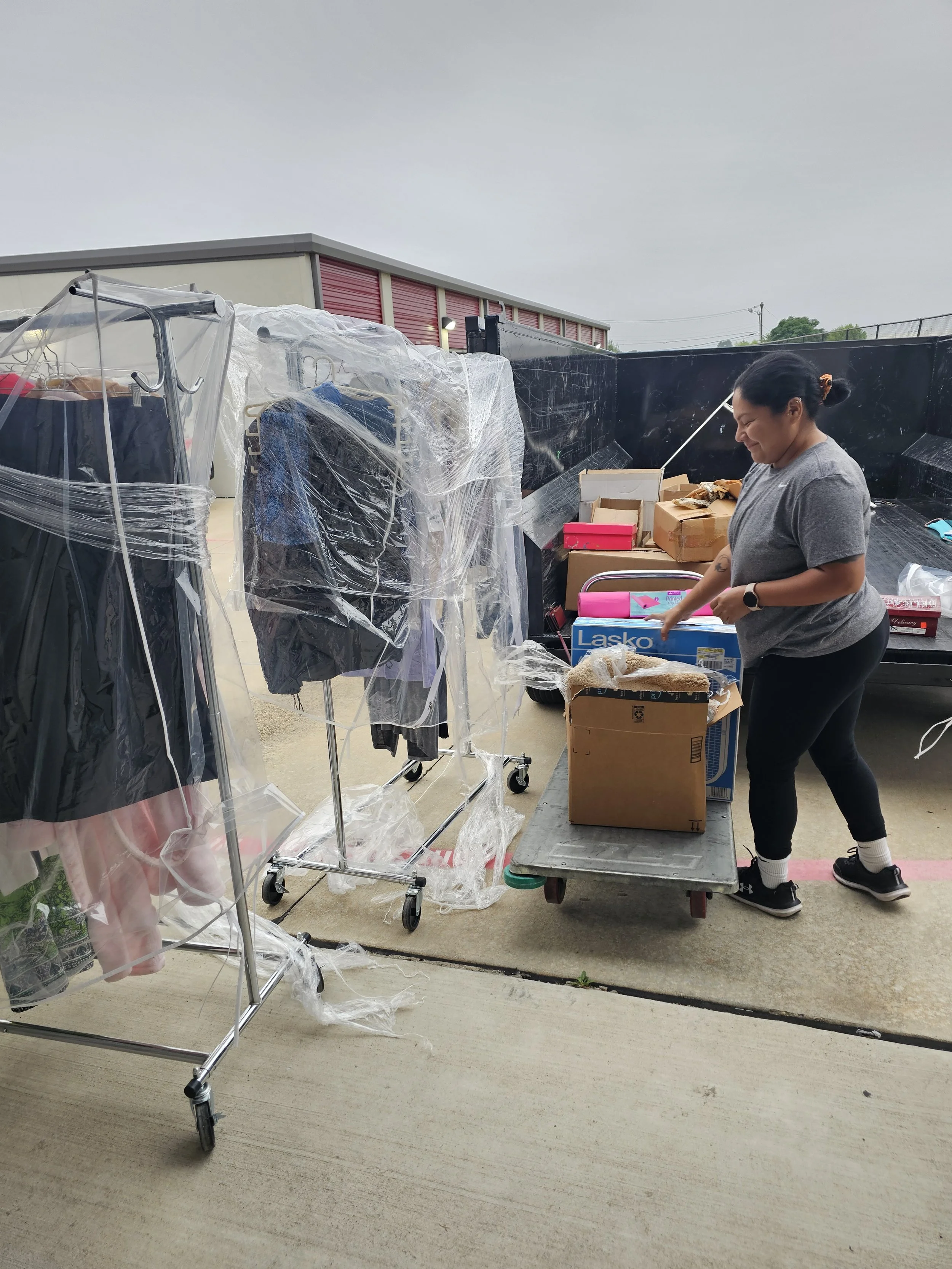 Woman organizing boxes on a cart outside, with clothes on a rack on the way to a donation center.