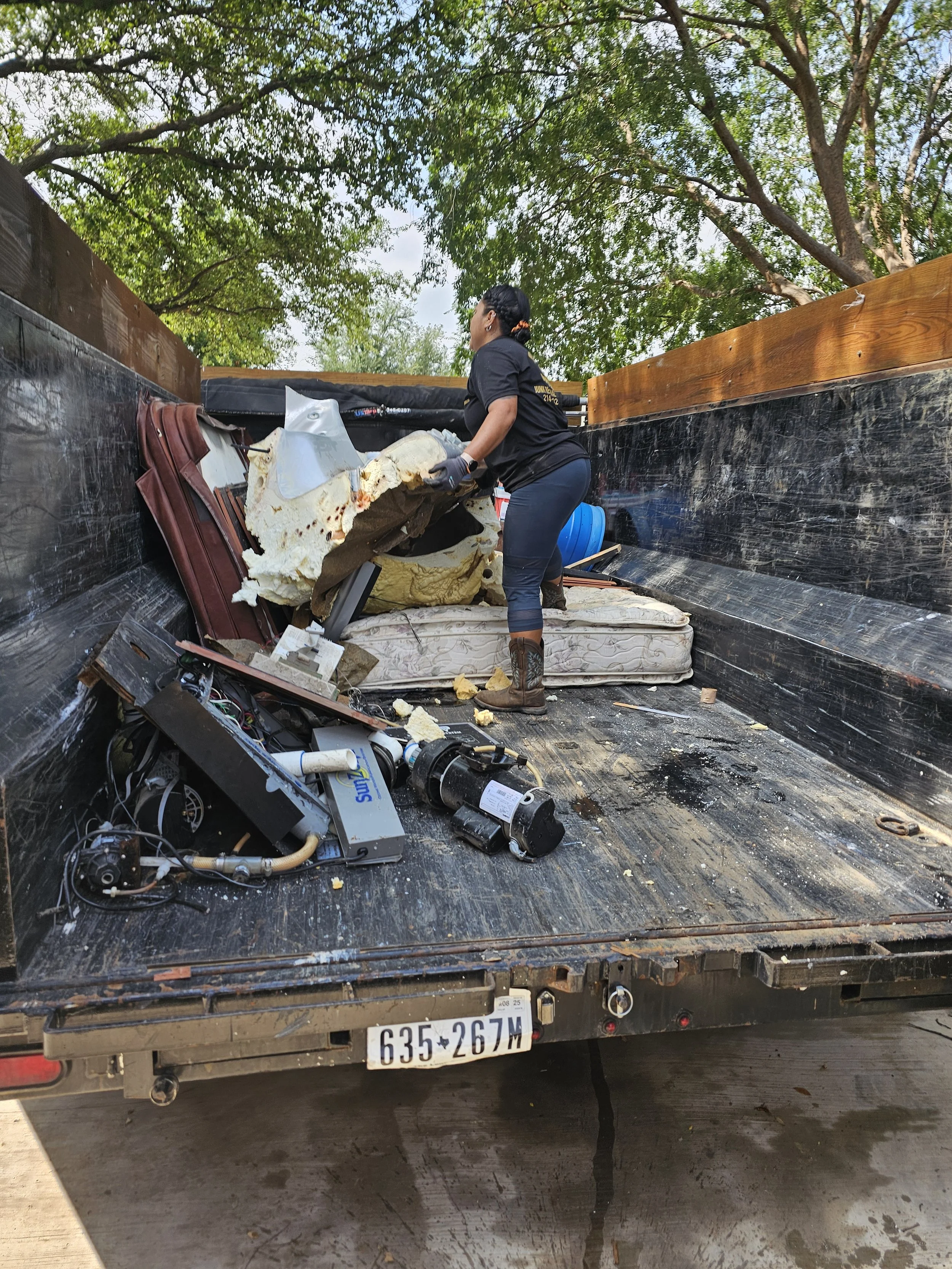 A woman is cleaning up debris and trash from the bed of a truck, which contains old furniture, insulation, and electronic equipment, with trees in the background.