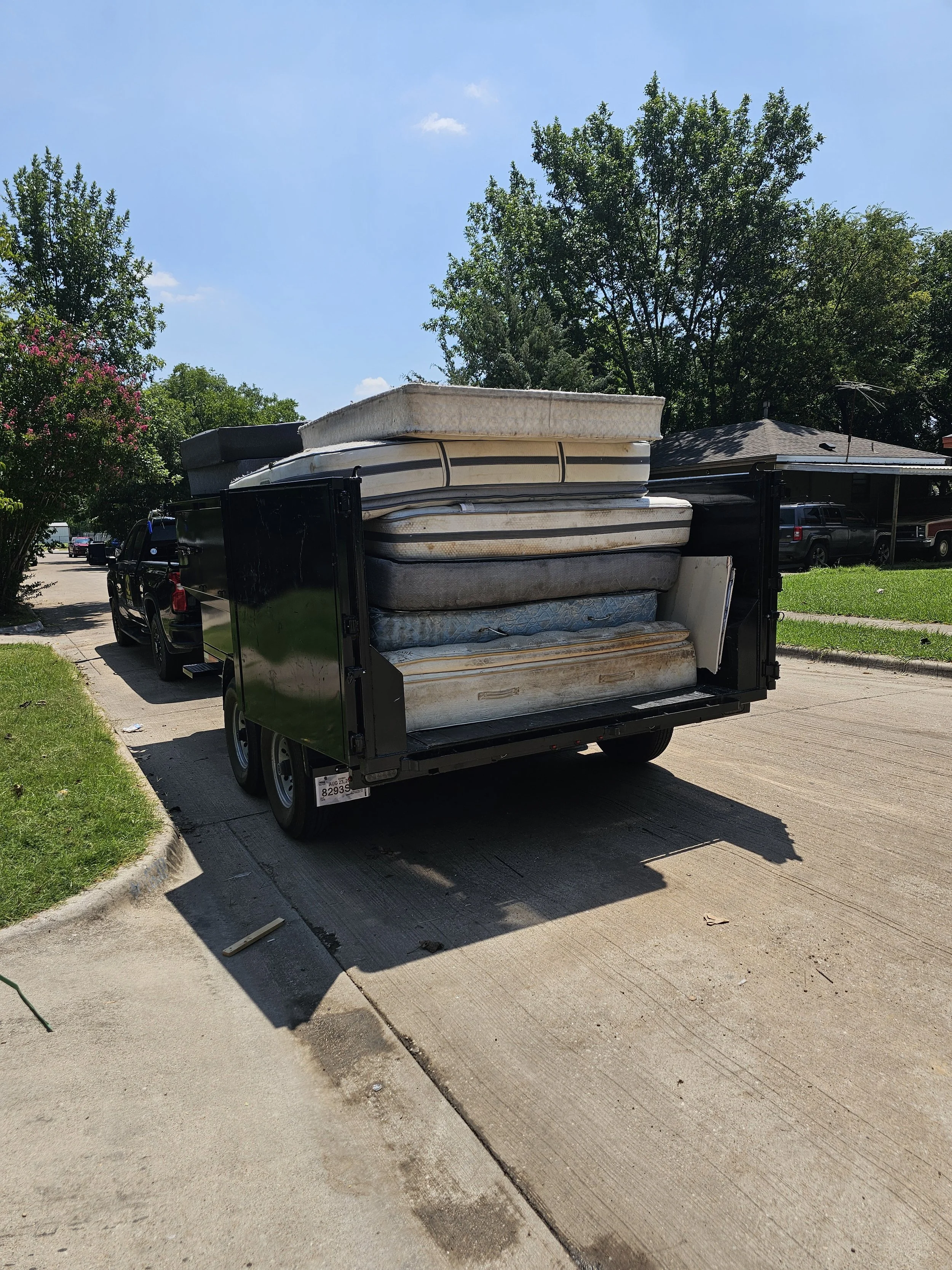 A black trailer attached to a truck, loaded with old mattresses and box springs, parked on a residential street under a sunny blue sky.