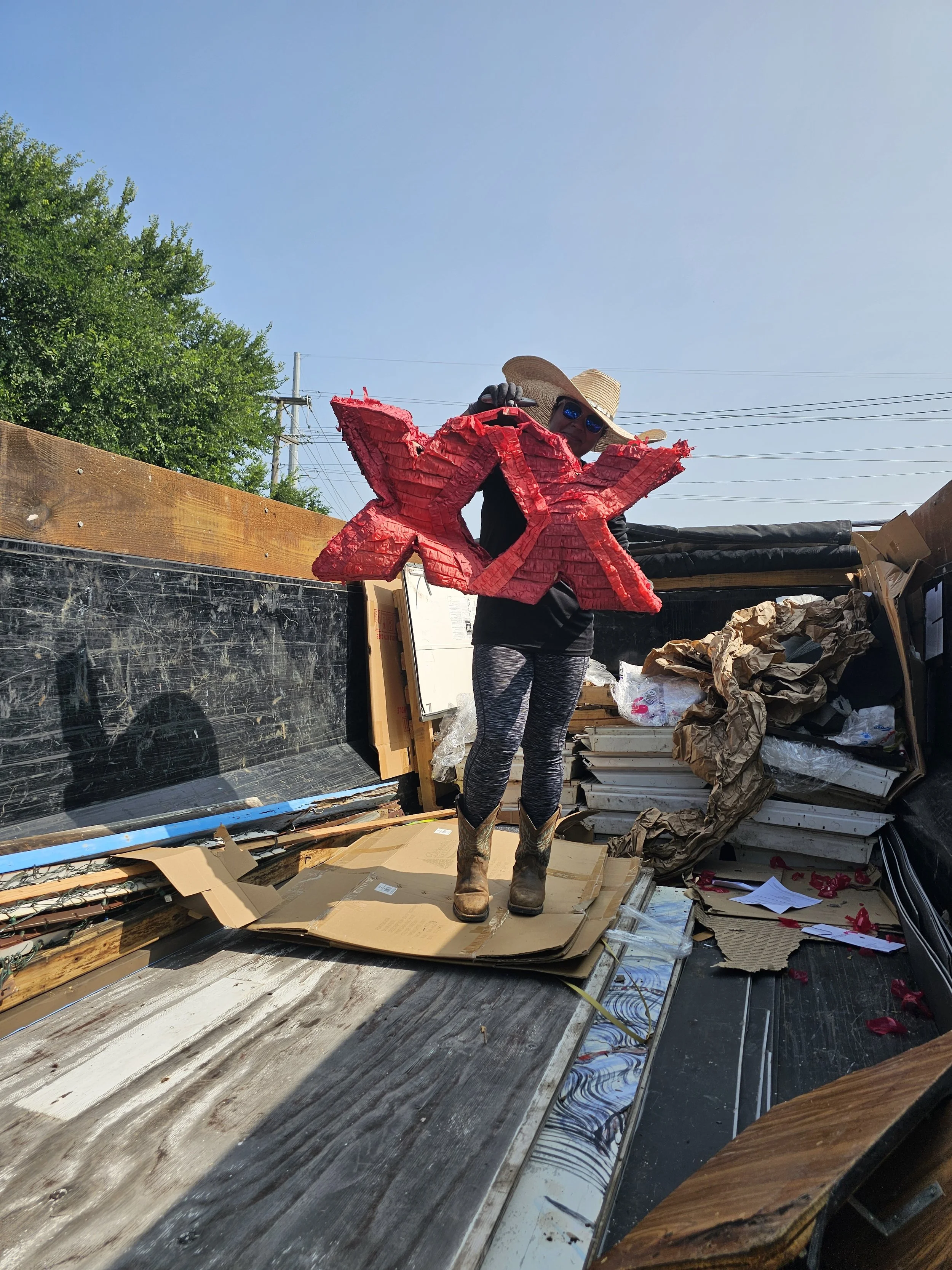 Person wearing cowboy boots, sunglasses, and a wide-brimmed hat holding a large red taco-shaped piñata in a truck bed filled with cardboard and paper trash on a sunny day with blue sky and green trees in the background.